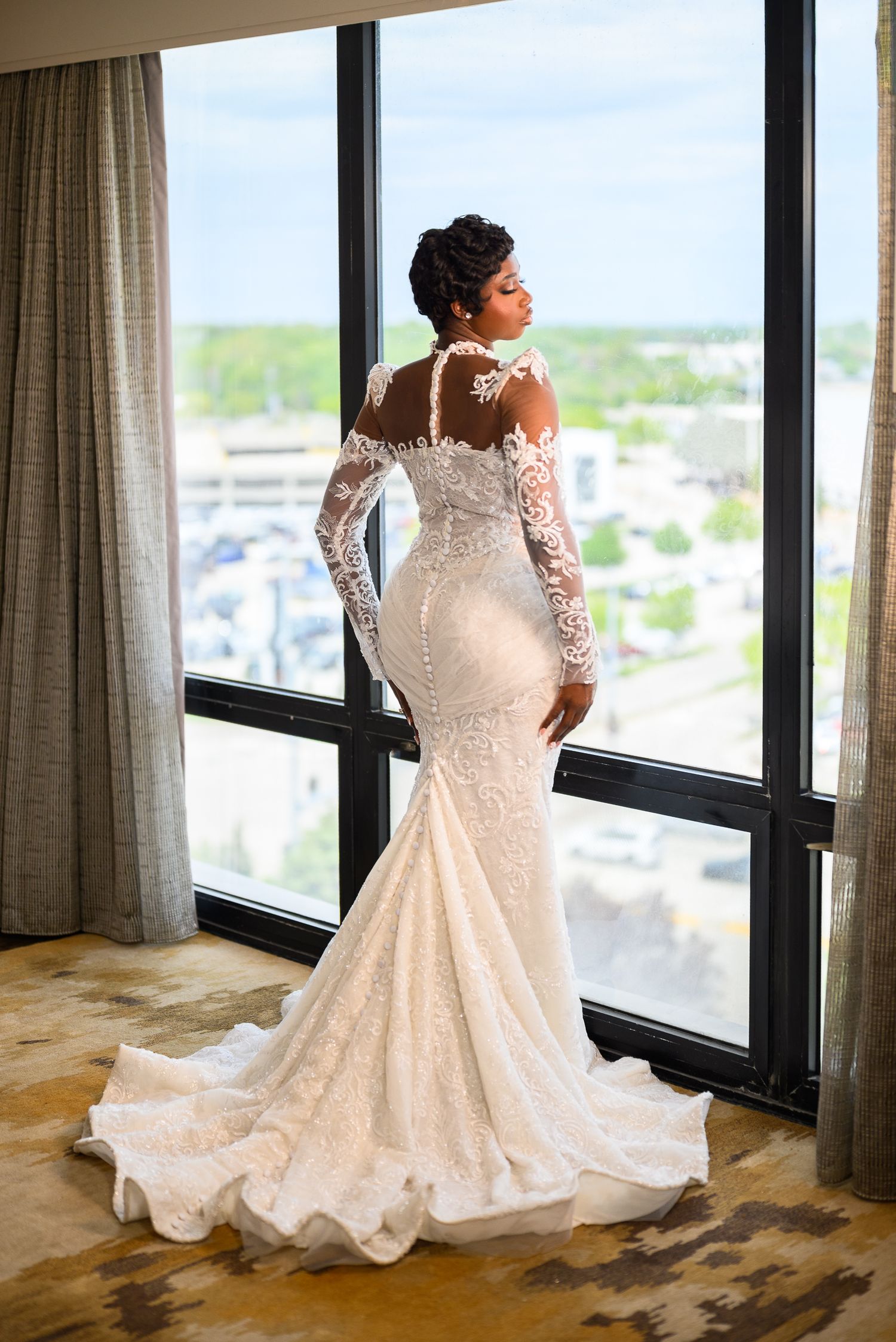 An elegant Black bride in a stunning lace mermaid wedding gown with long sleeves stands by floor-to-ceiling windows.