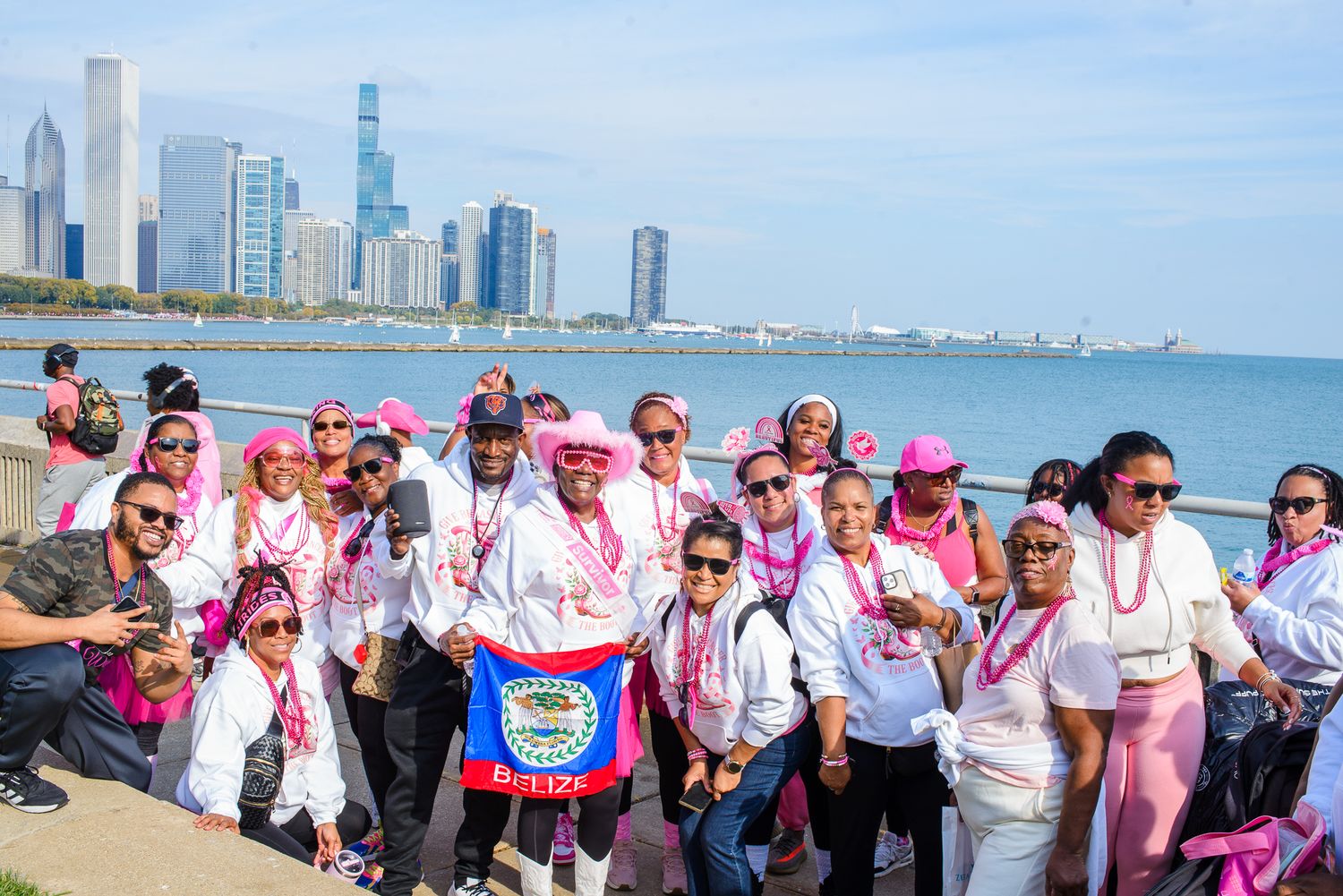 A large group wearing pink attire and sunglasses poses together along Chicago's lakefront with the city skyline in view.