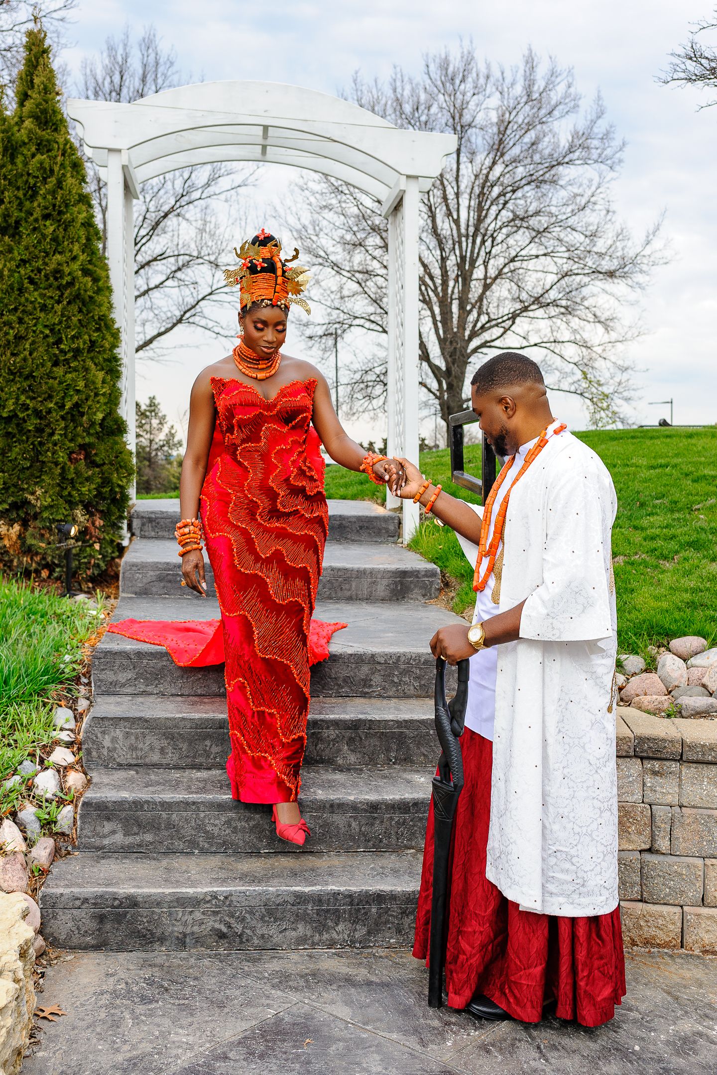 A groom in white traditional attire helps his bride in a vibrant red gown descend stone steps at an outdoor ceremony.