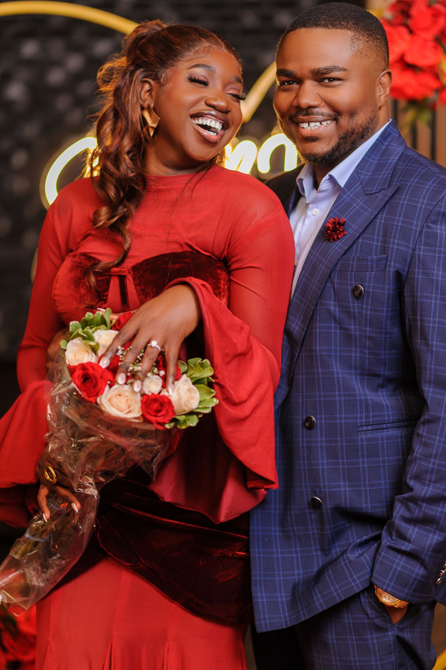 Couple smiling at a romantic proposal, woman in red dress holding roses, man in blue plaid suit.