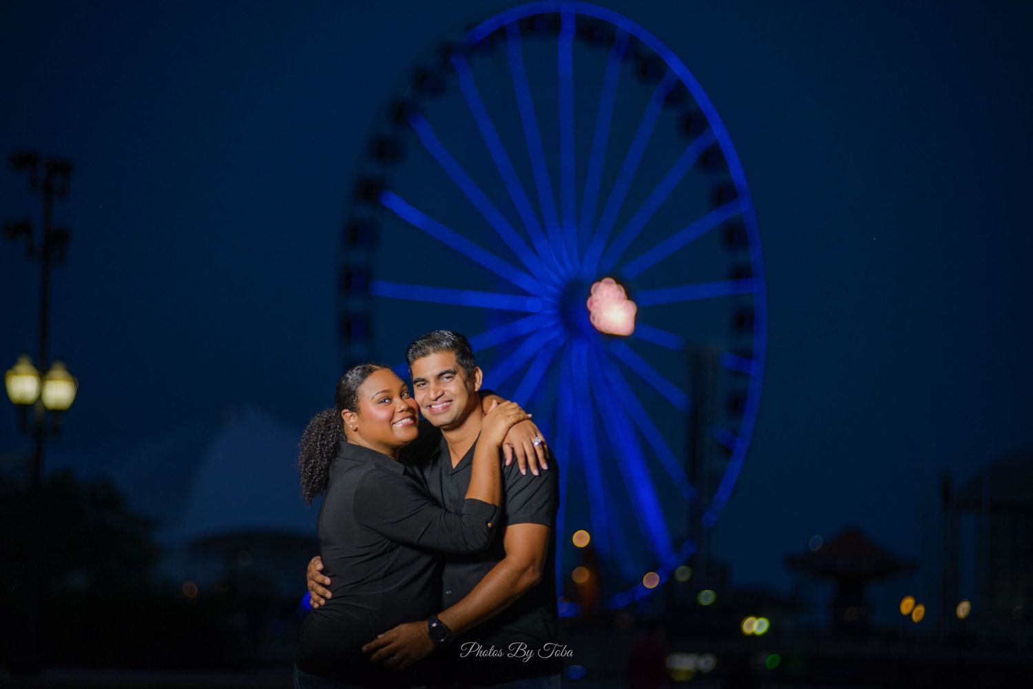 A couple shares an intimate moment at night with a glowing blue ferris wheel illuminated in the background.