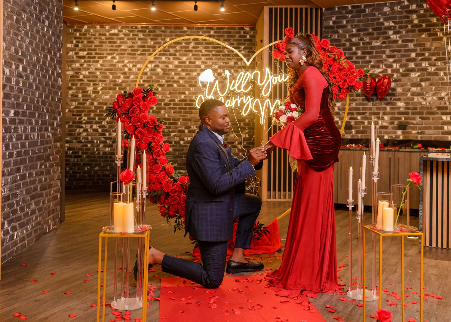 Man proposing to woman in red dress at romantic setup with heart arch, red roses, and Will You Marry Me neon sign.