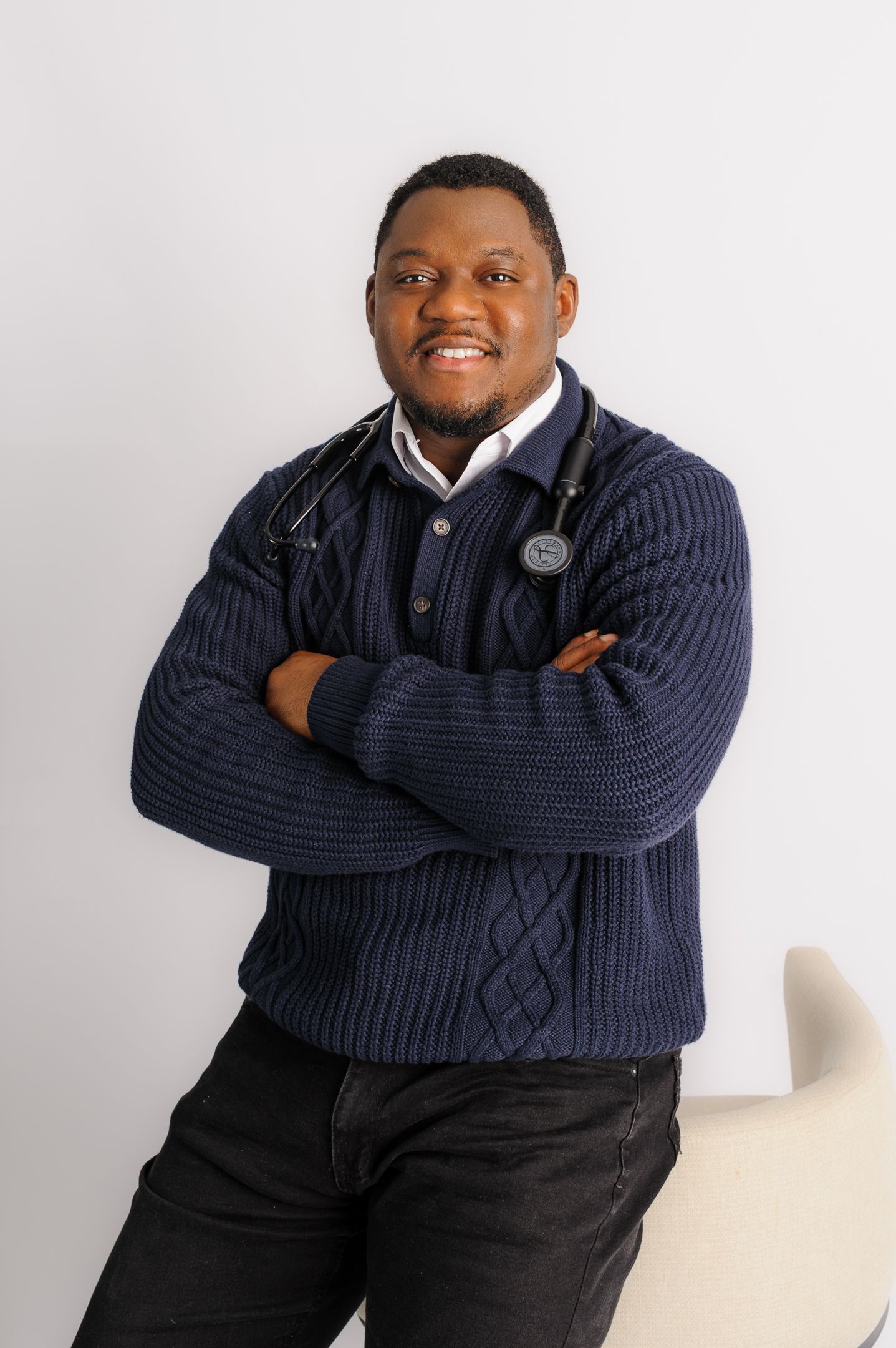 Smiling male doctor in navy sweater with stethoscope, arms crossed, posing confidently against white background.