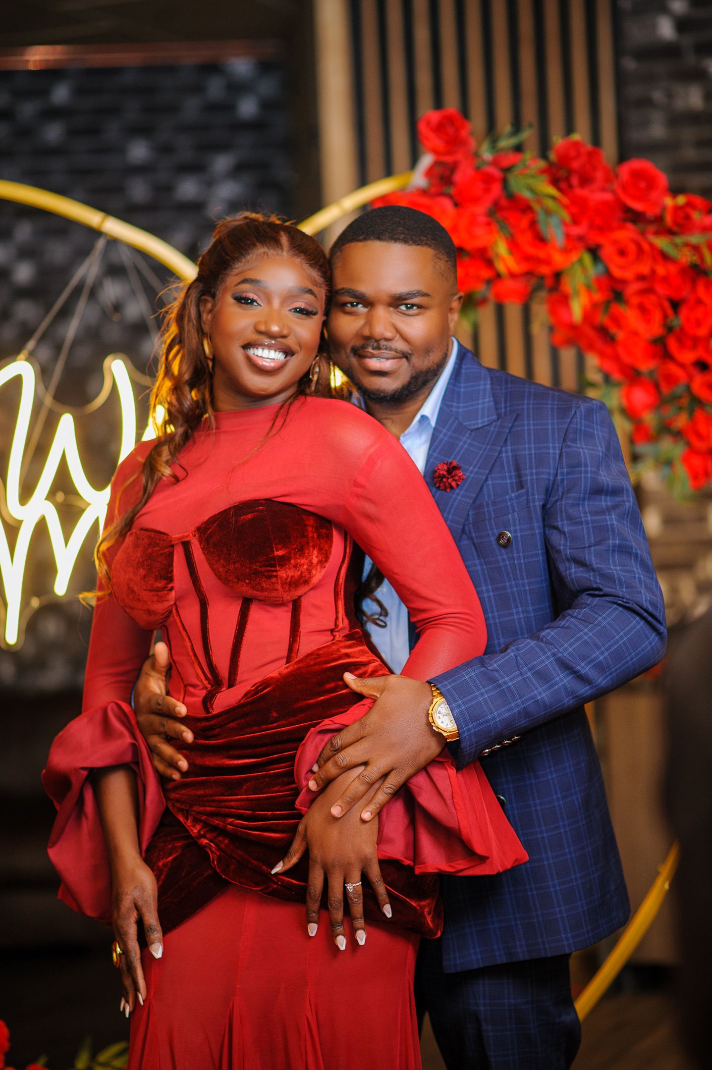 Couple posing elegantly, woman in red velvet gown and man in blue plaid suit, with red roses backdrop.