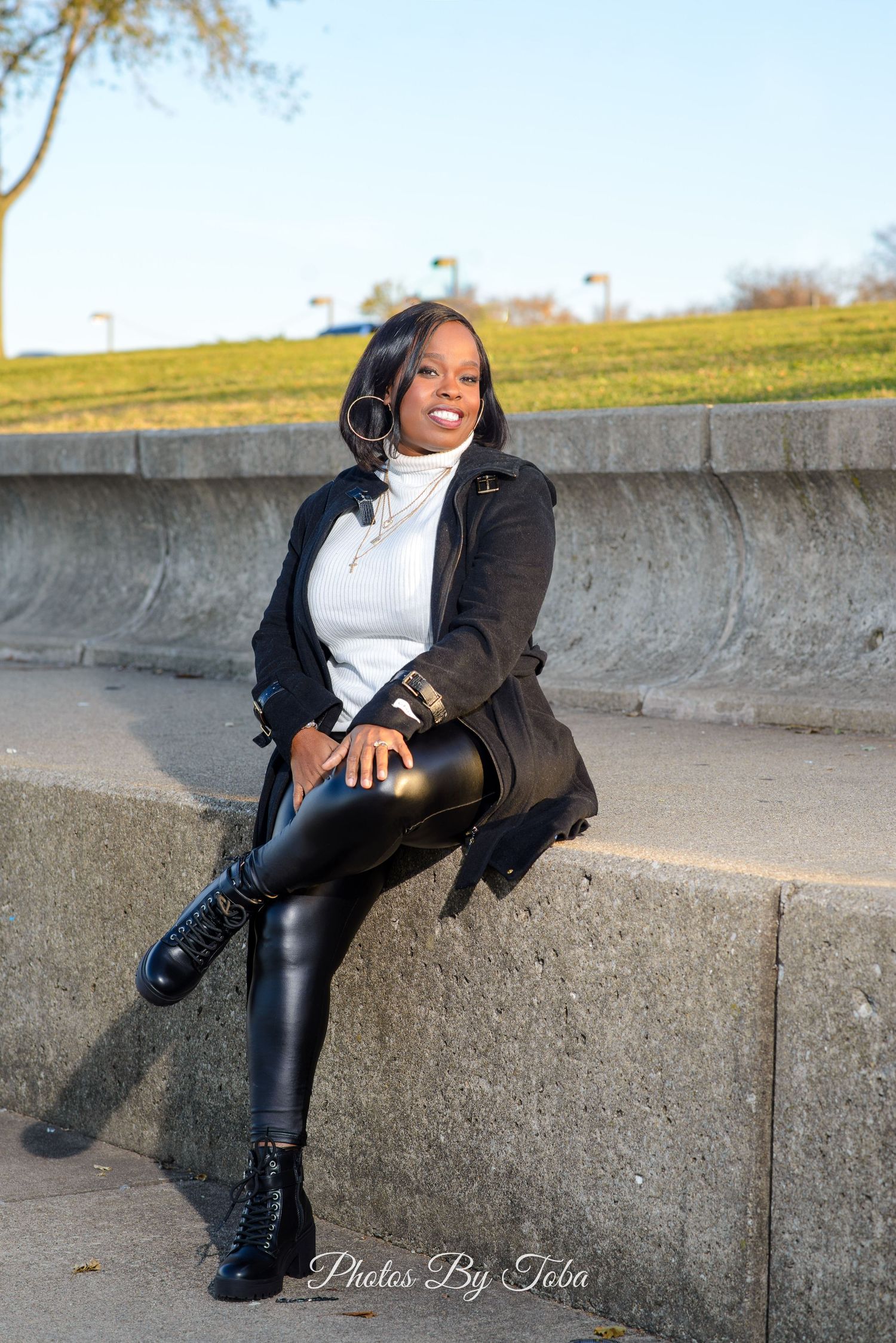 Someone wearing a brown jacket, white top and leather pants poses casually on a concrete wall outdoors.