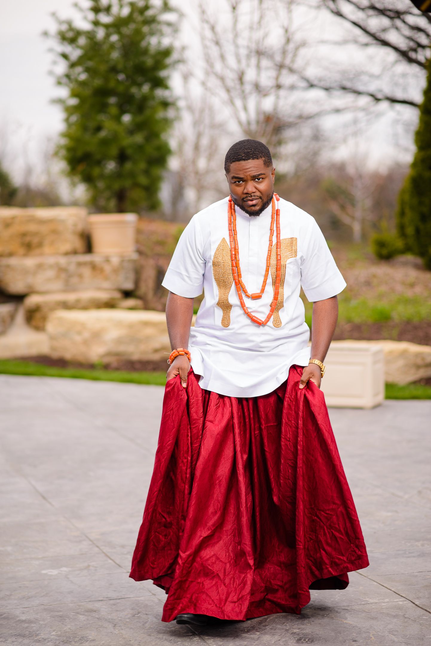 Nigerian groom in white agbada and red wrapper with orange coral beaded necklace at outdoor wedding.