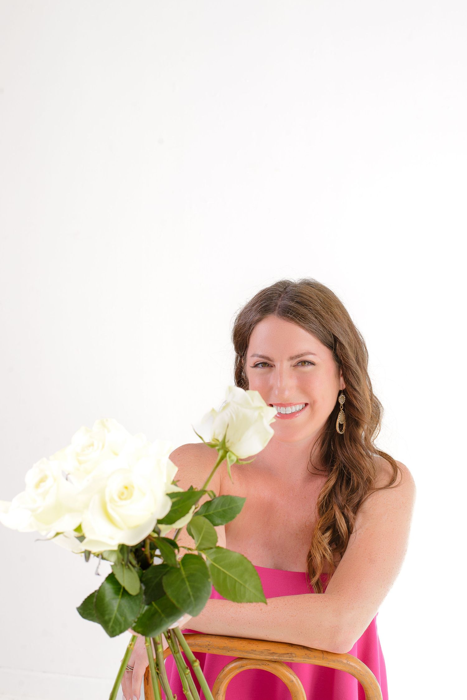 Smiling woman in pink dress holding white roses bouquet, seated on wooden chair against white background.