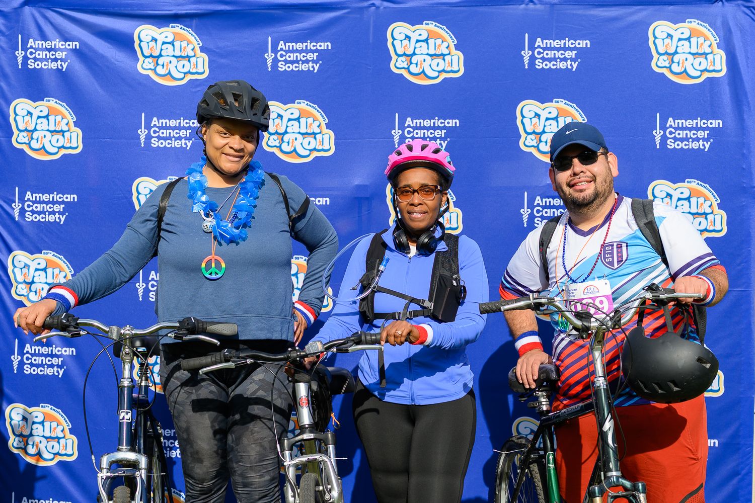 Three cyclists with bikes pose together at American Cancer Society Walk & Roll Chicago event backdrop.