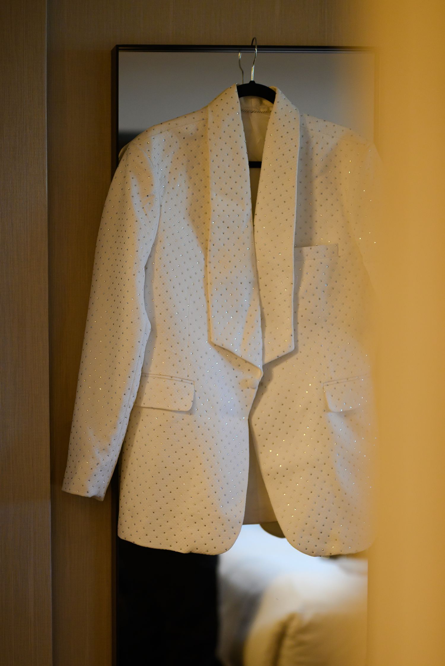 White embellished blazer jacket hanging on a hanger against a mirror in a hotel room.