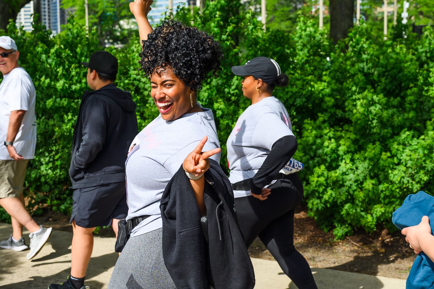 Laughing woman in white event shirt walking energetically past green hedges at outdoor race.