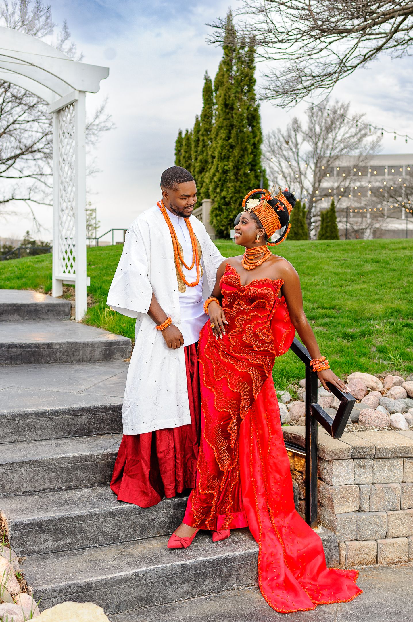 Couple in vibrant Nigerian traditional wedding attire, man in white agbada, woman in red and orange lace gown, posing on steps.