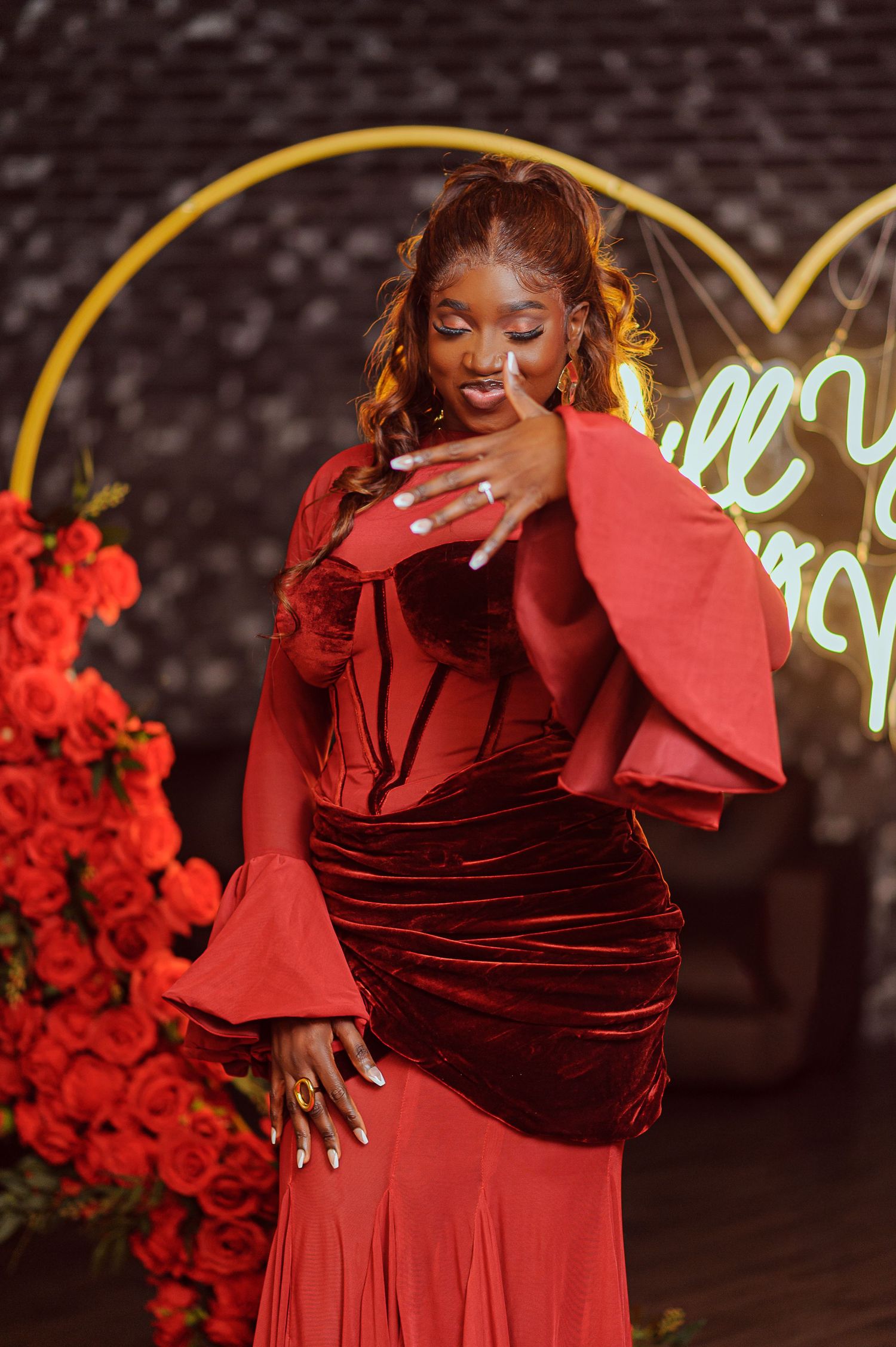 Woman in red velvet gown poses elegantly before heart-shaped neon sign and rose arrangement.