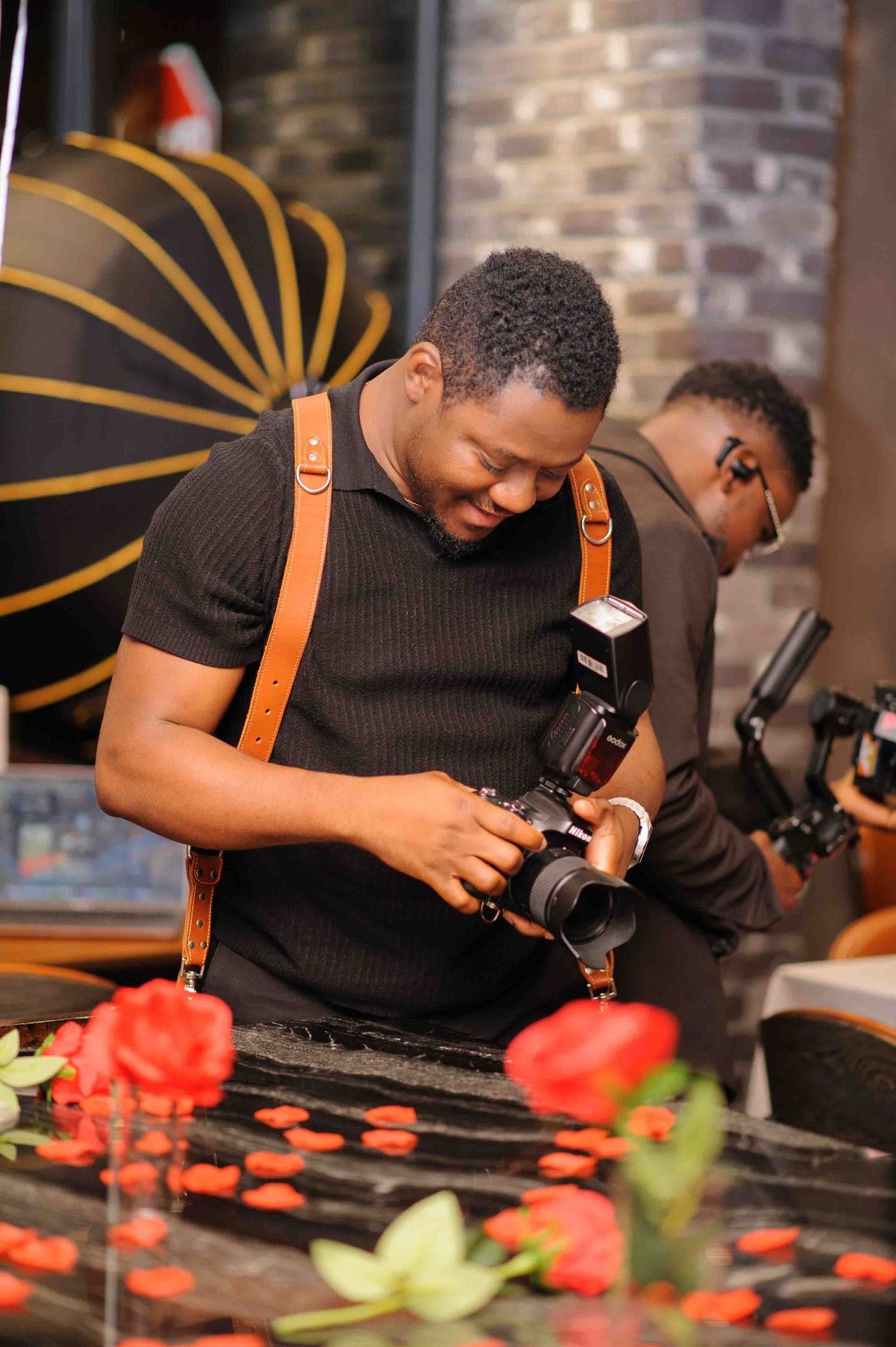 A smiling photographer reviews shots on his camera while wearing a leather strap at an event with red roses.