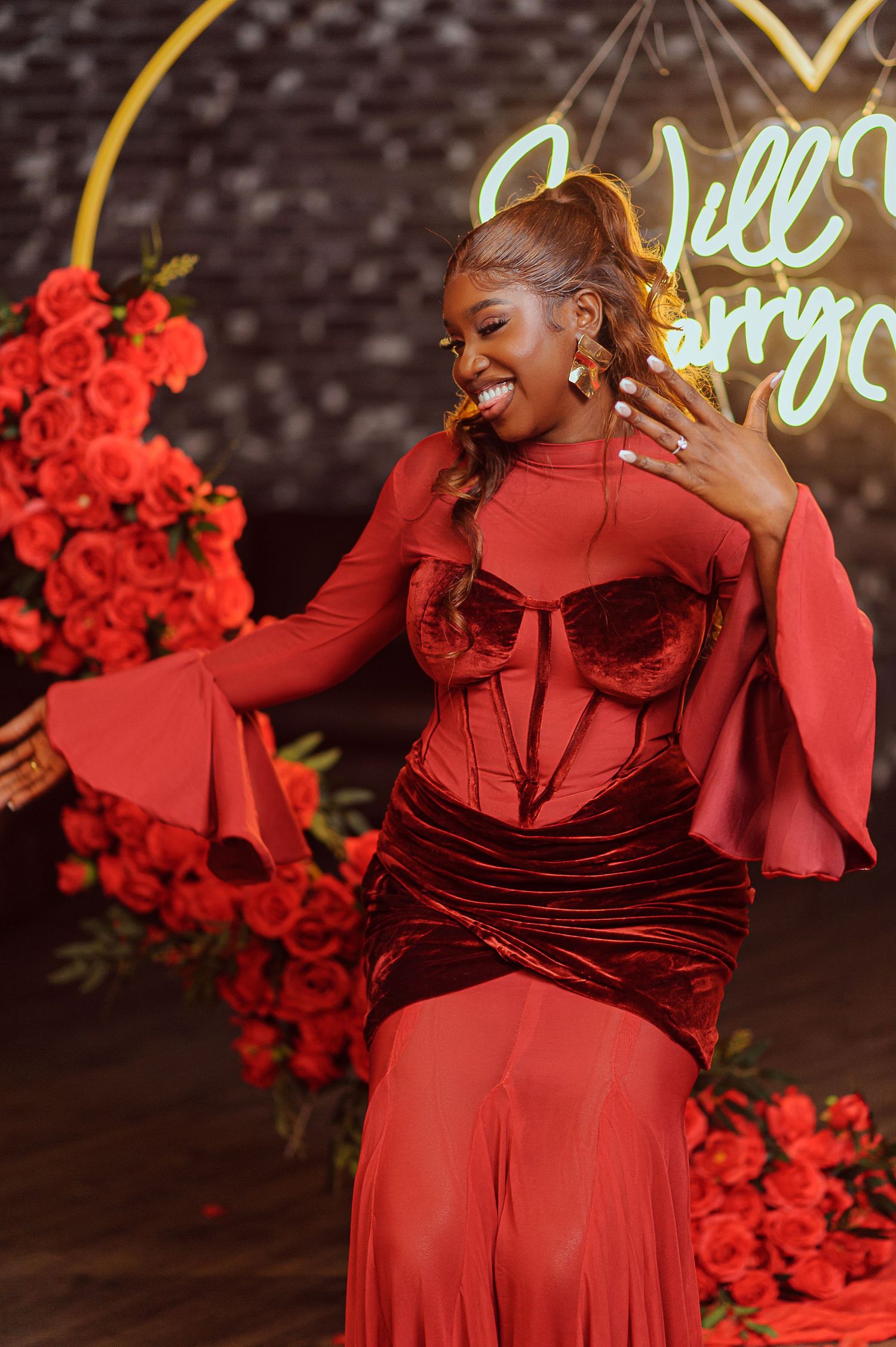 Woman in red corset gown smiling at engagement proposal setup with roses and neon sign.