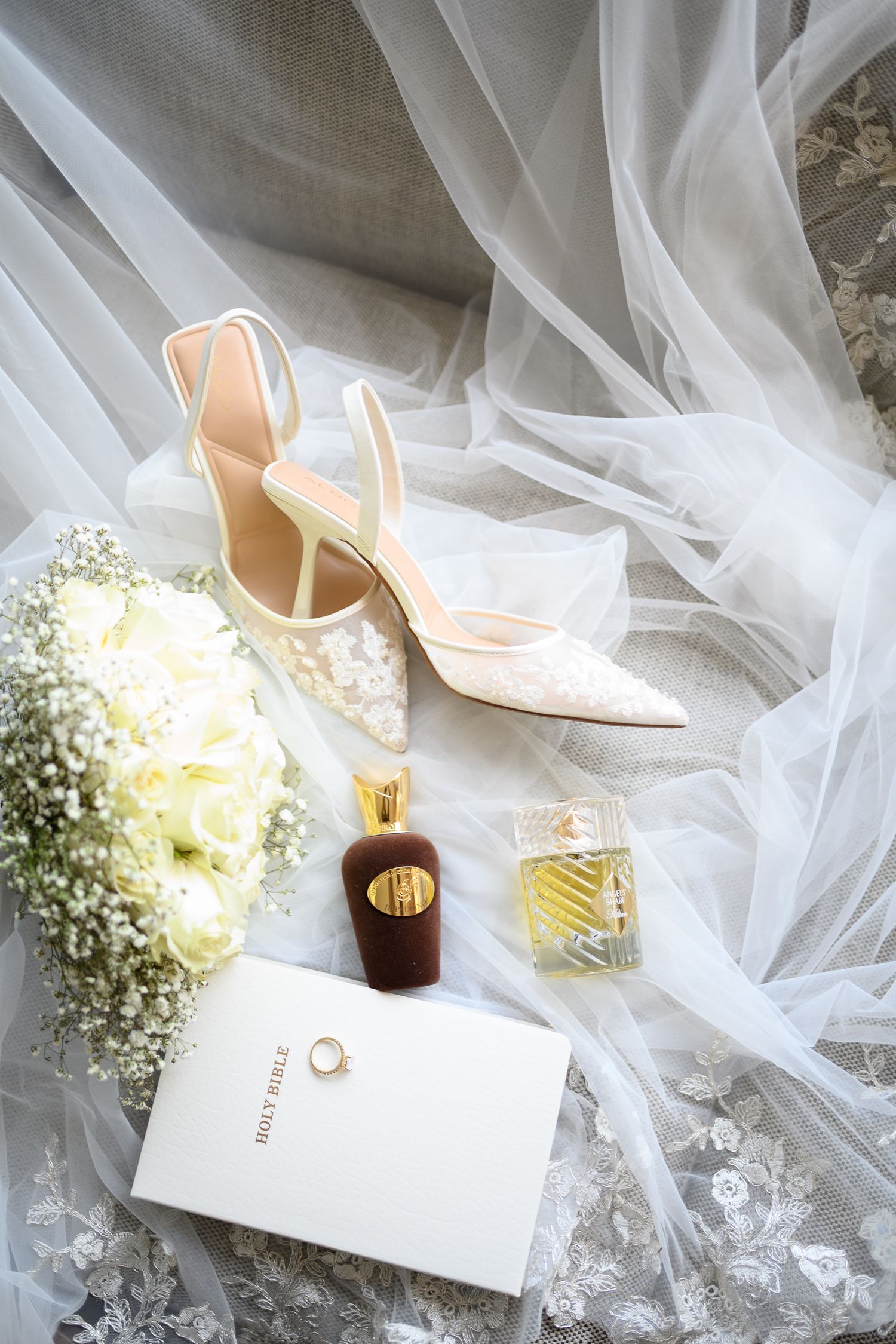 Bridal flat lay with lace heels, white rose bouquet, perfume bottles, gold ring, and Holy Bible on sheer veil fabric.