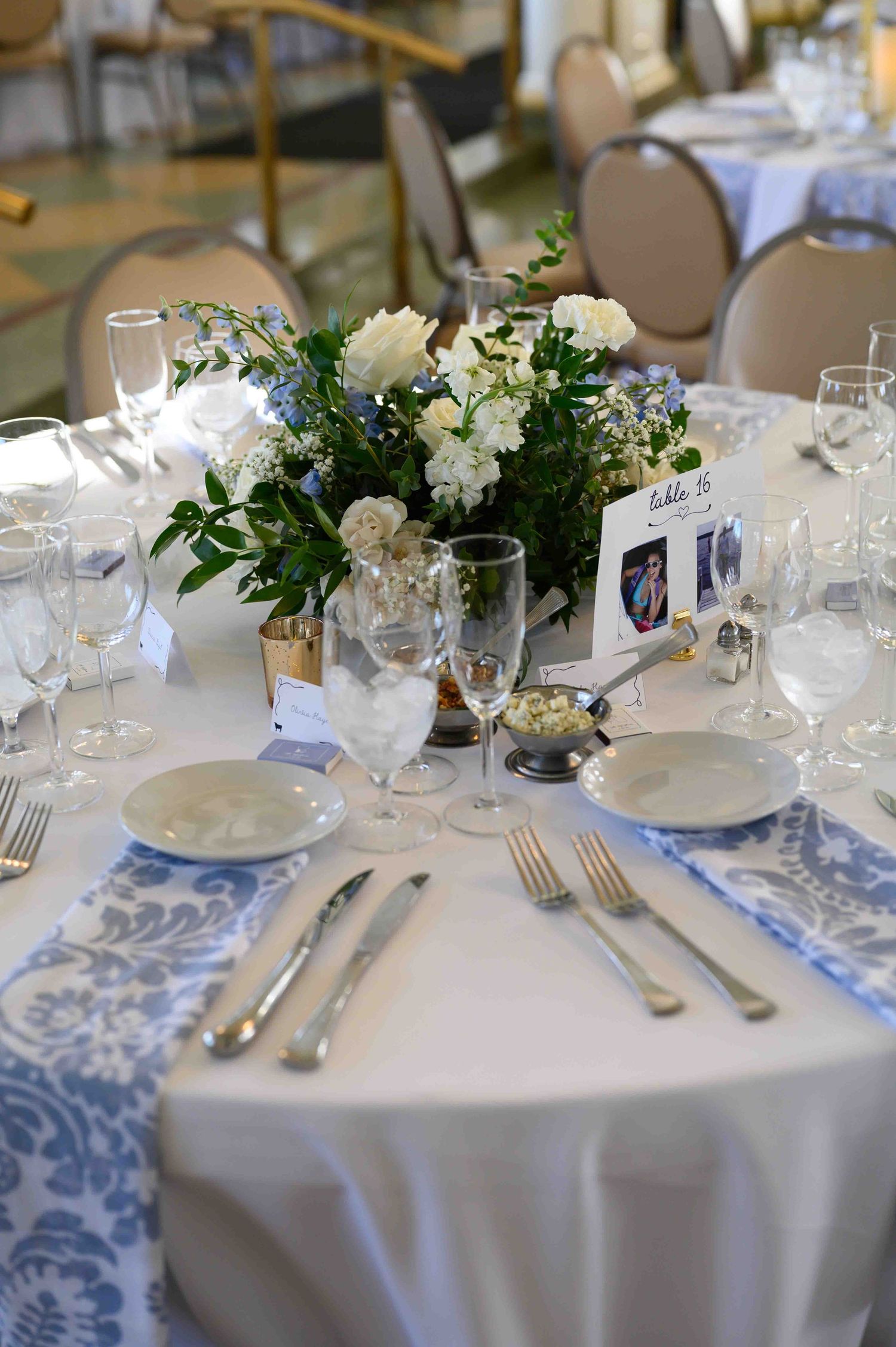Elegant wedding reception table set with white linens, blue damask runner, crystal glasses, silver cutlery, and white floral centerpiece.