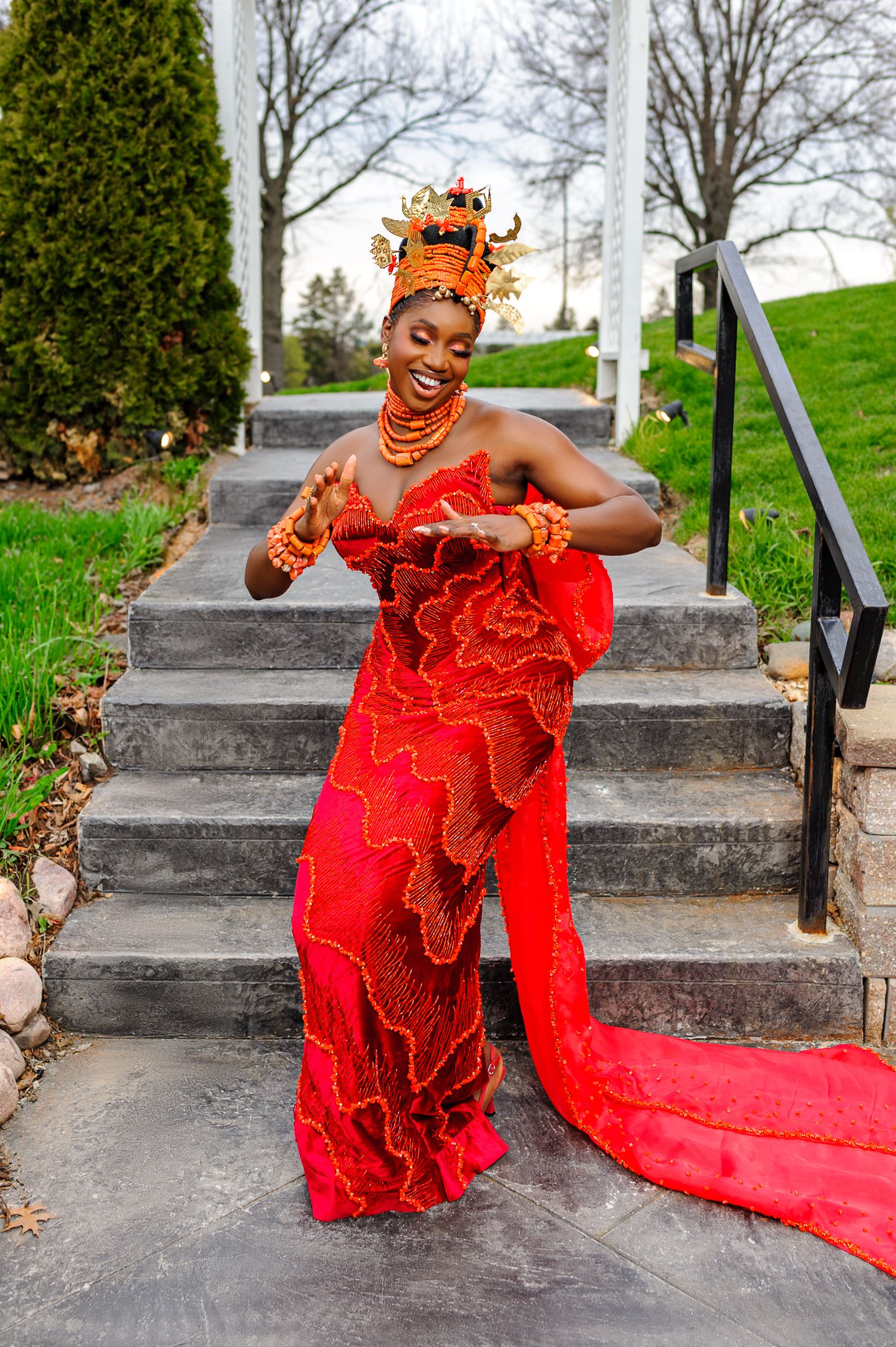 Joyful Nigerian bride in red beaded traditional attire laughs on outdoor stone staircase.