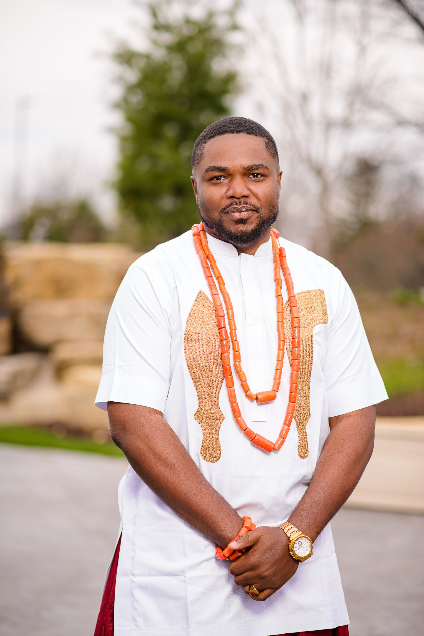 Man in white Nigerian traditional agbada shirt adorned with orange coral bead necklaces at outdoor venue.