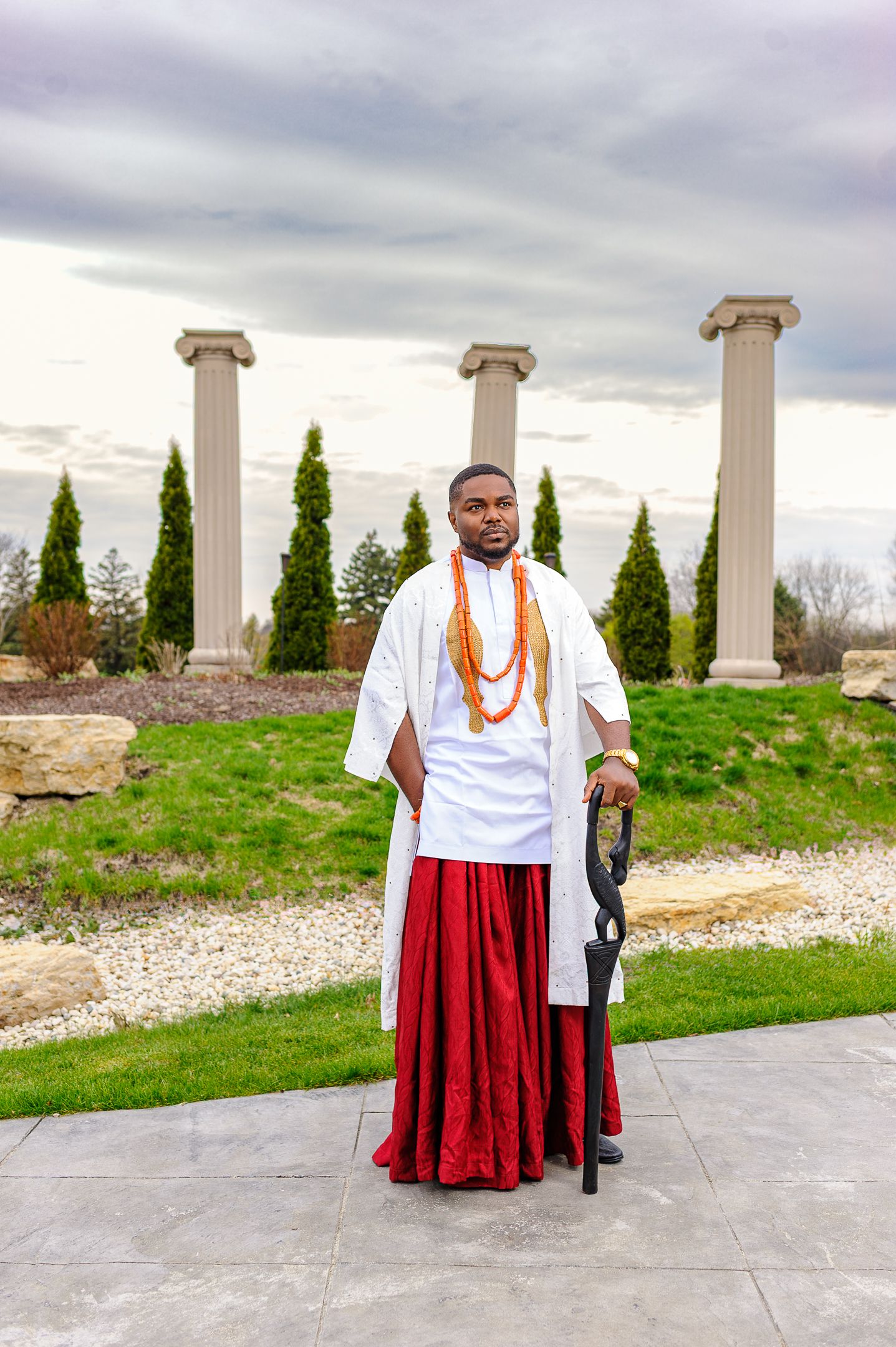 Man in white and red Nigerian traditional outfit with coral beads holding a ceremonial staff outdoors.