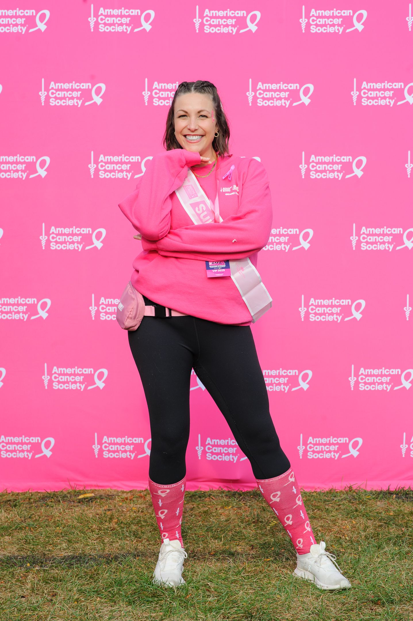Woman in pink sweatshirt strikes playful pose at cancer awareness event with branded backdrop.