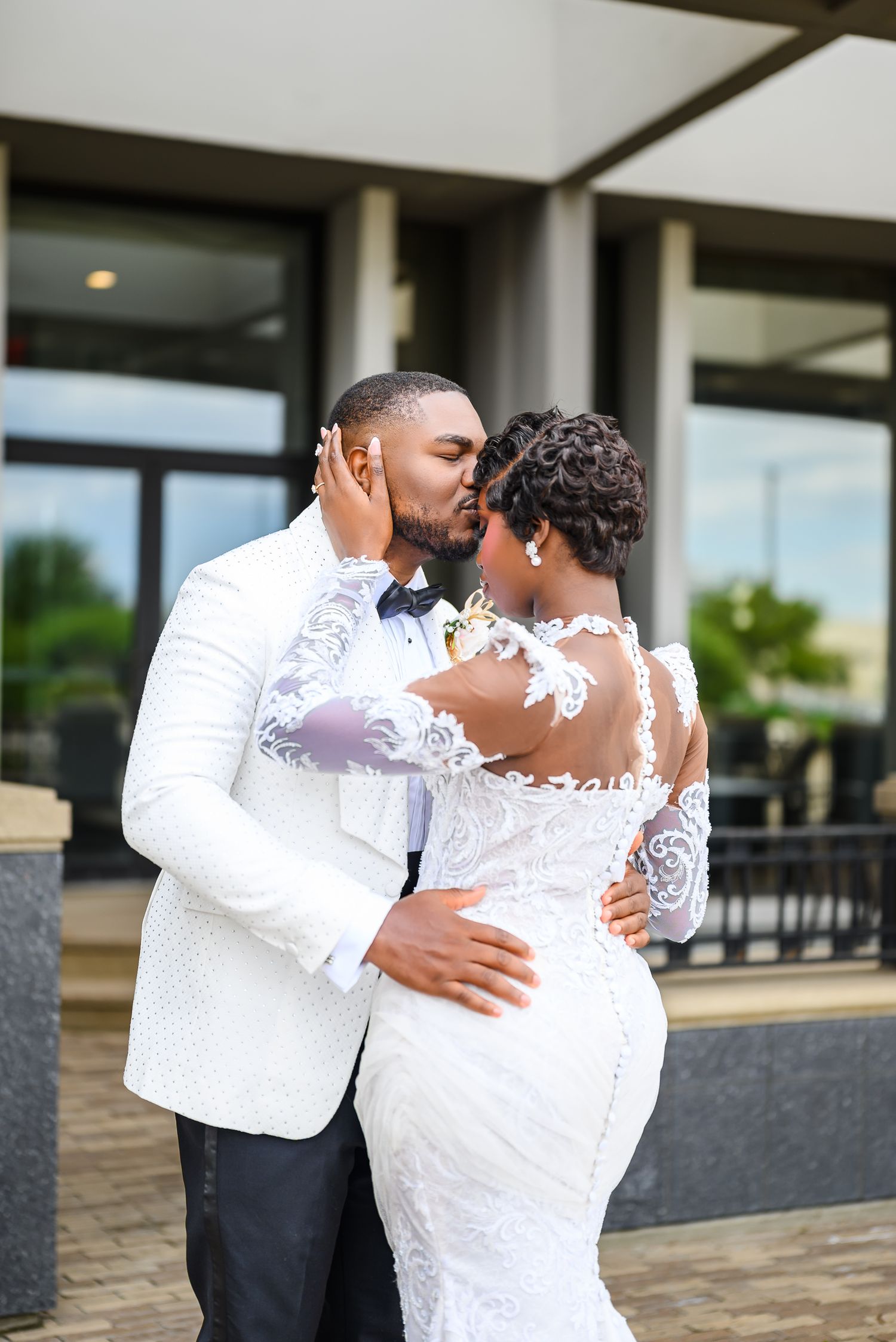 A bride and groom share a romantic kiss outdoors, dressed in elegant white wedding attire with lace details.