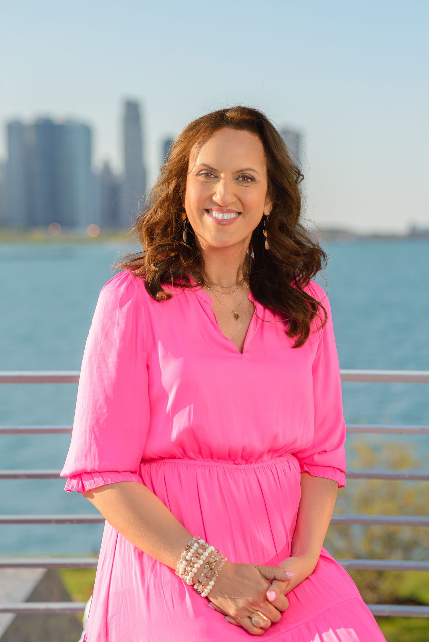 Person in bright pink dress poses on waterfront deck with city skyline in background on sunny day.