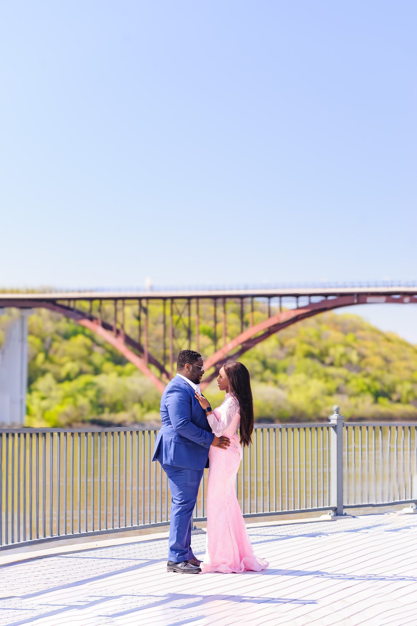 Elegant couple in blue suit and pink gown embracing on a waterfront deck with a red arch bridge in background.