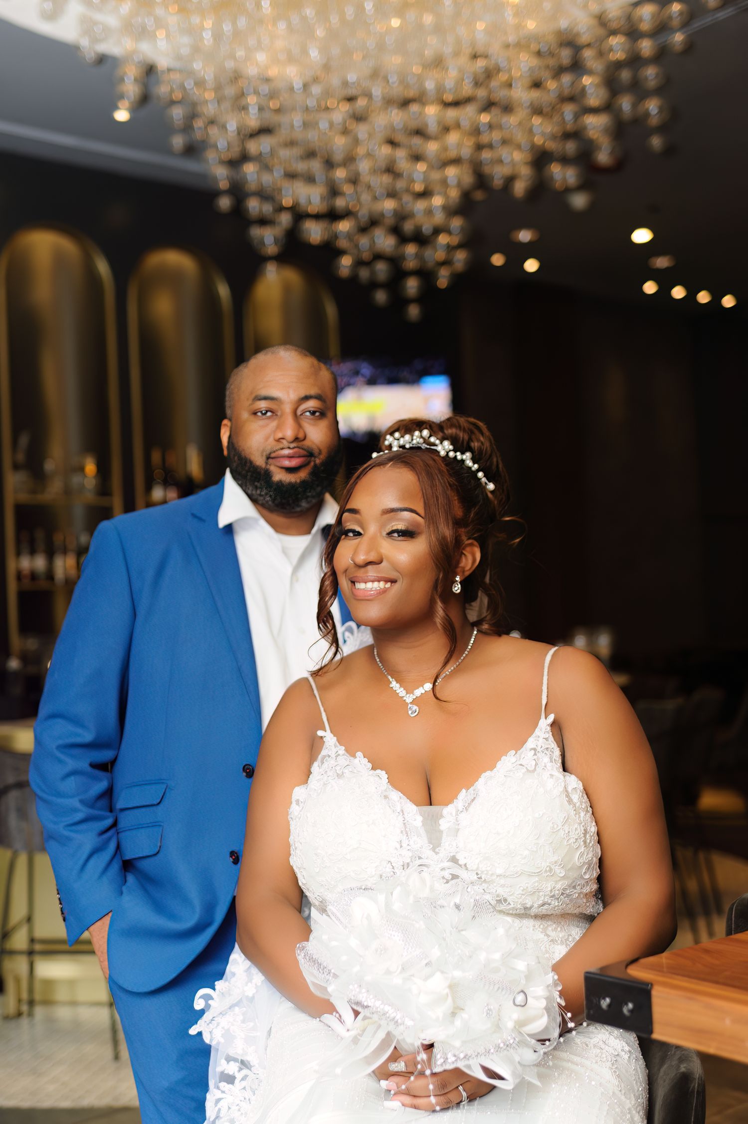 Bride in white lace gown and groom in blue suit pose elegantly under crystal chandelier at wedding reception.