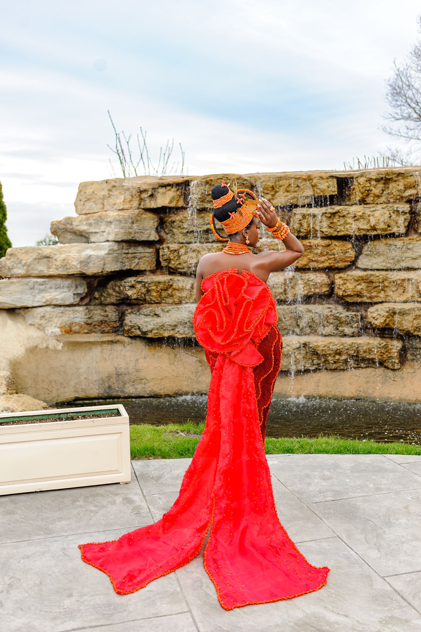 Woman in red Nigerian traditional bridal gown with long train posing by stone waterfall feature outdoors.