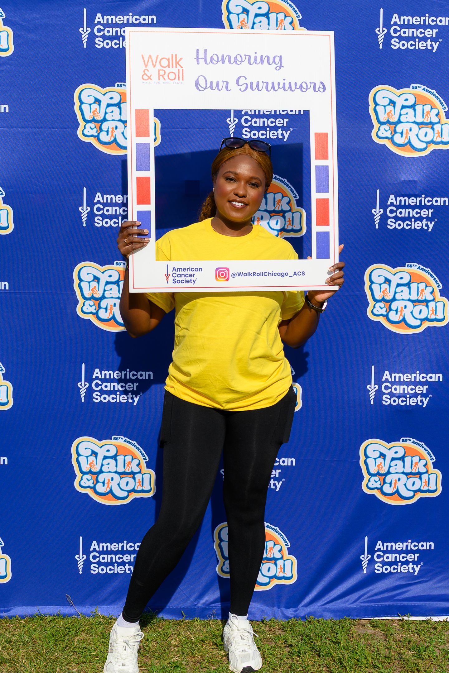 Woman in yellow shirt holds Walk & Roll American Cancer Society frame at outdoor cancer survivor celebration event.