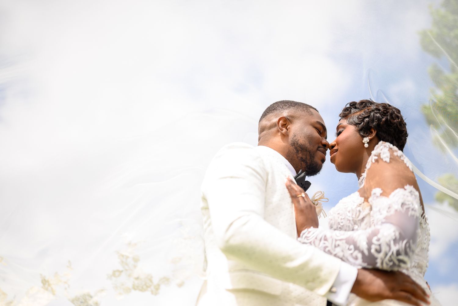 A Black bride and groom share a romantic kiss outdoors, with the bride's lace veil flowing in the breeze.