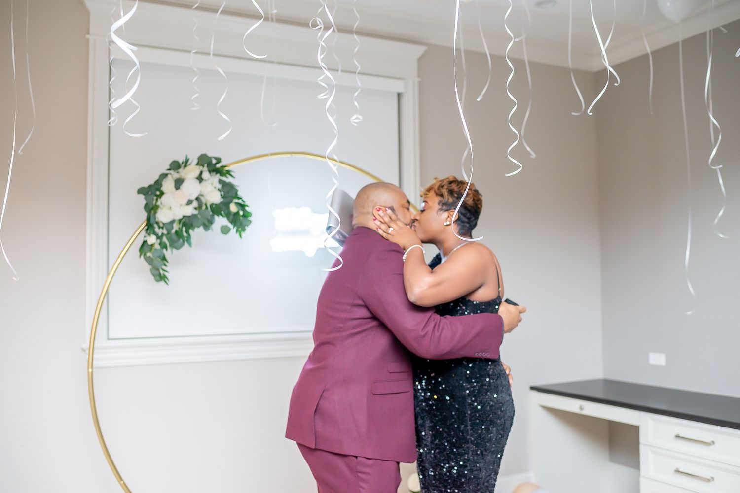 Couple sharing first kiss under gold hoop arch with greenery, confetti falling, in decorated room with silver streamers.