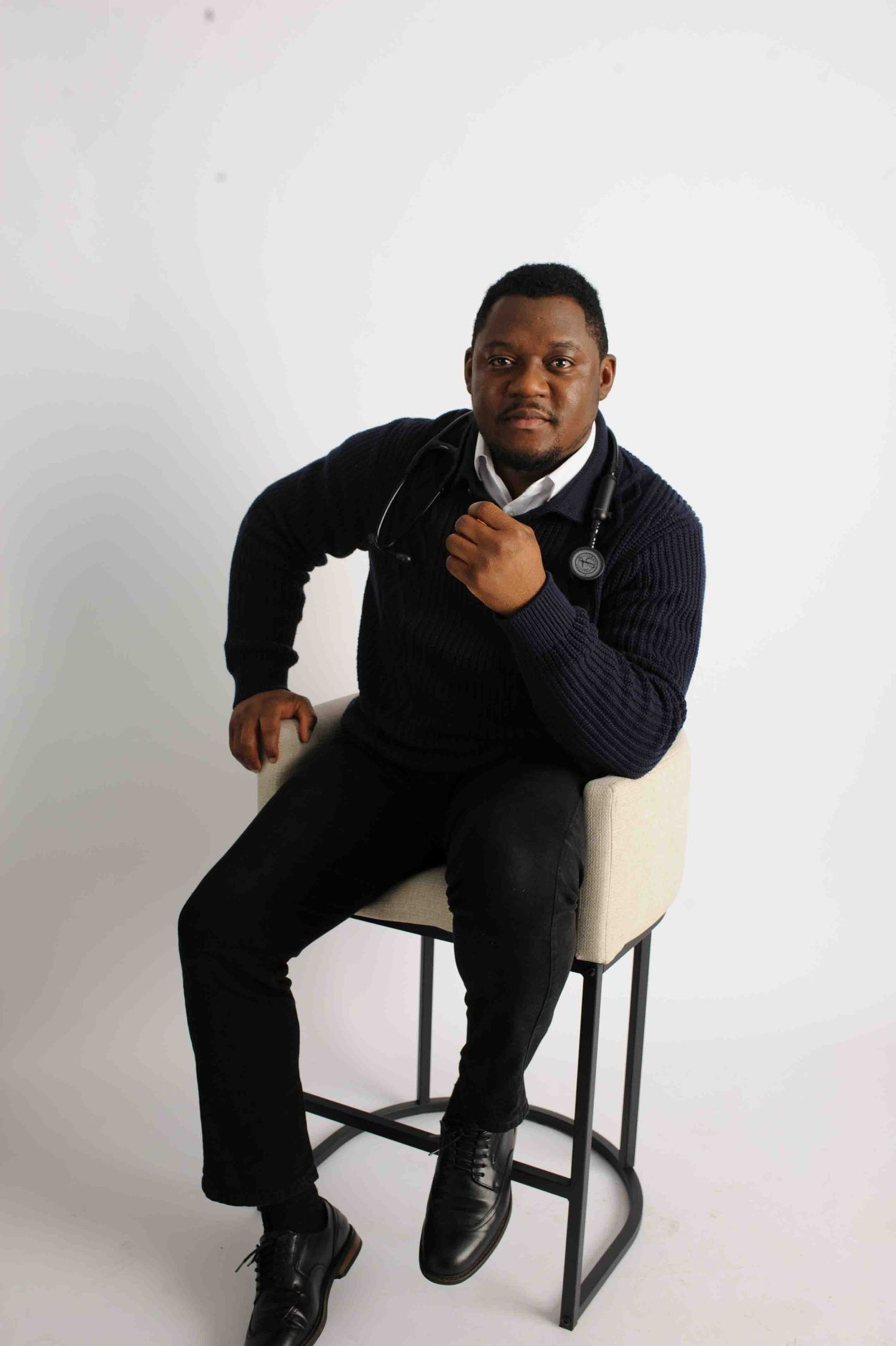 Medical professional in navy sweater with stethoscope seated on stool against white background.