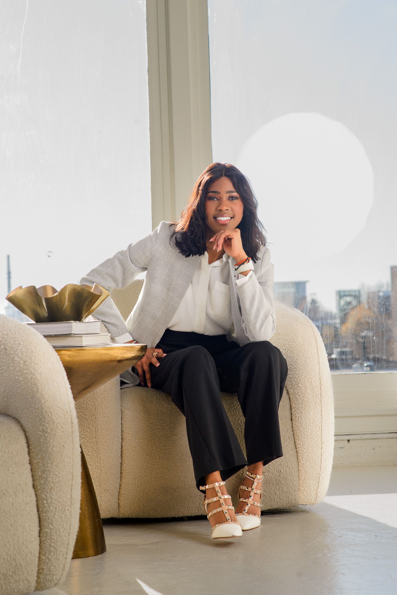 Professional woman in gray blazer and black pants sitting in a modern white chair by large windows.