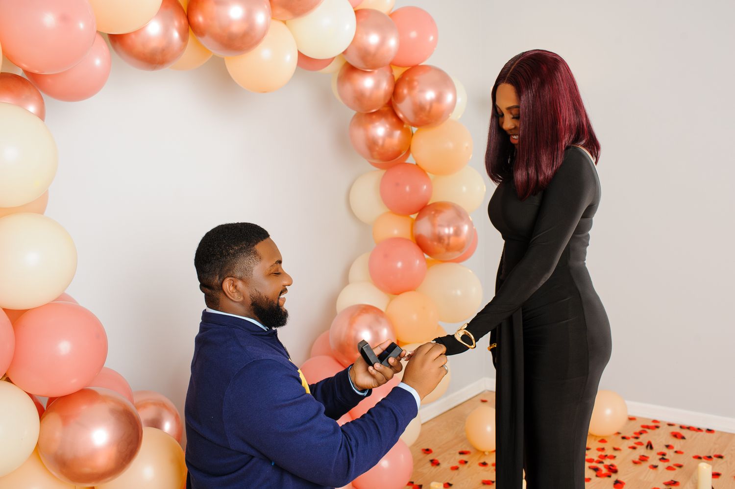 Man proposes on one knee to woman in black outfit, surrounded by pink and gold balloon arch with rose petals.