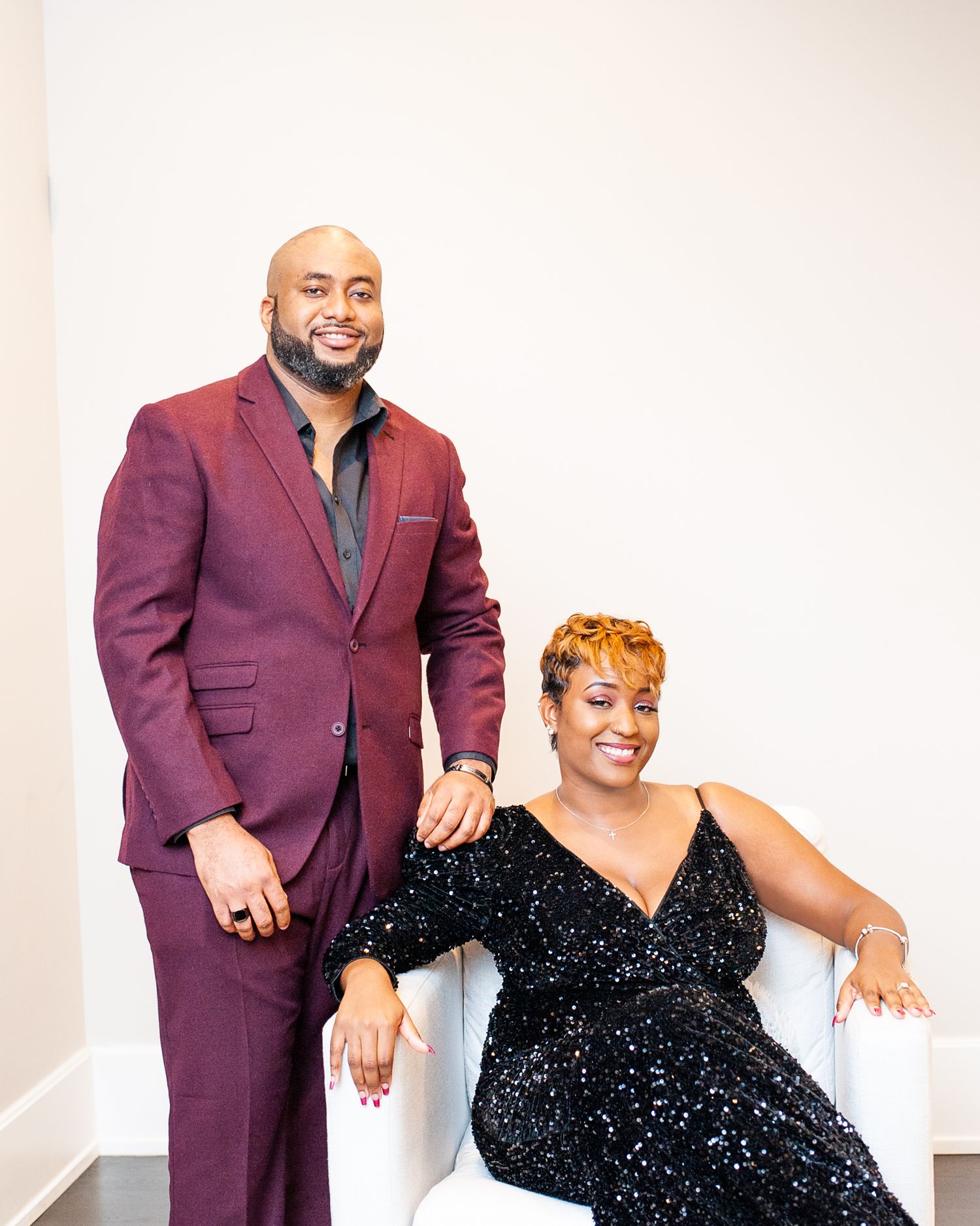 A man in a burgundy suit stands beside a woman in a black sequin dress seated in a white chair, posing elegantly.