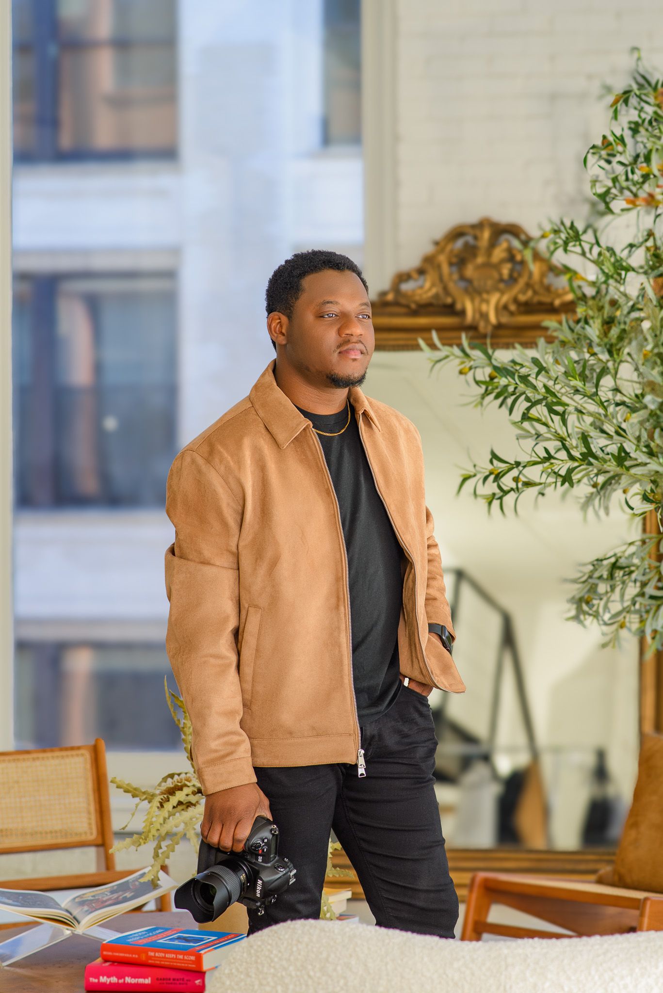 A young Black man in a tan jacket holds a camera while standing in a stylish modern interior space.