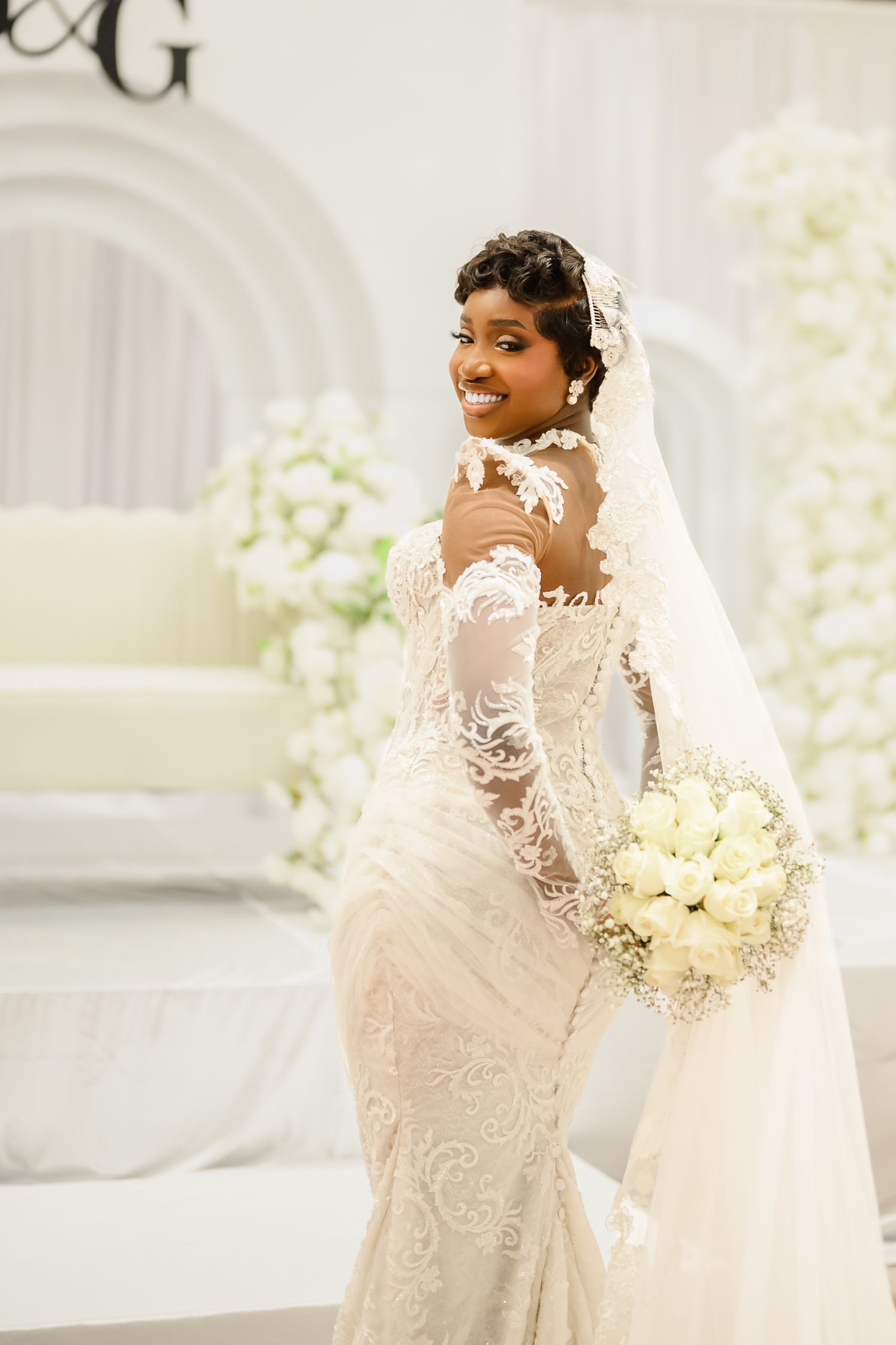 Smiling Black bride in lace mermaid gown and veil holds white rose bouquet in elegant white wedding venue.