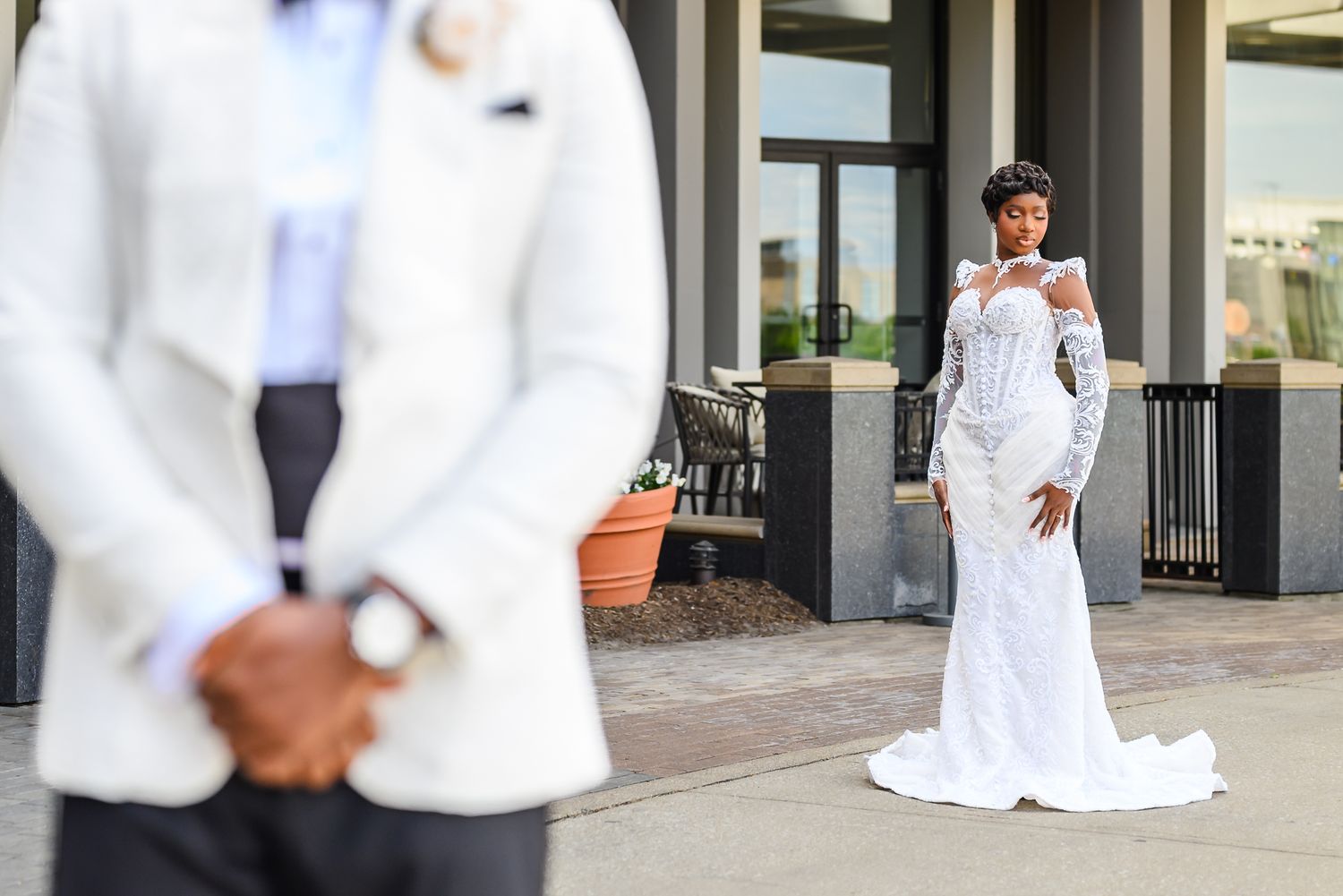 Bride in white lace mermaid gown stands behind groom in white suit during outdoor wedding photo session.