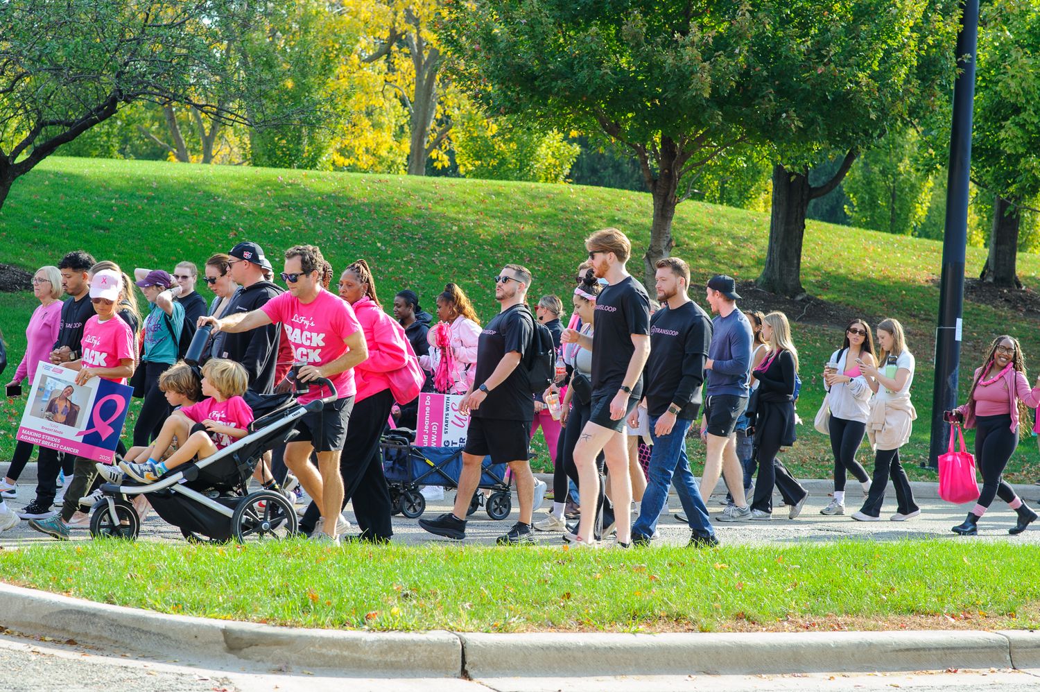 A large group of people walking together in a line along a scenic park path on a sunny day.