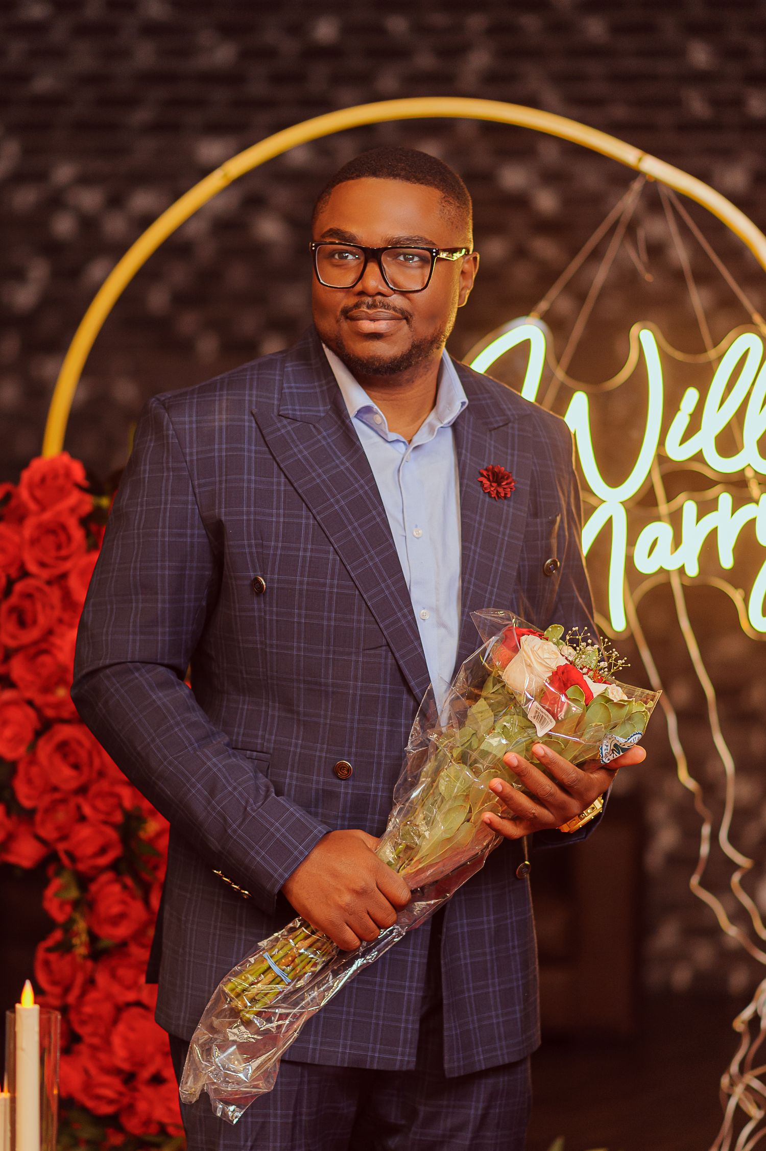 Well-dressed man in plaid blue suit holding bouquet of roses at elegant event with neon sign backdrop.