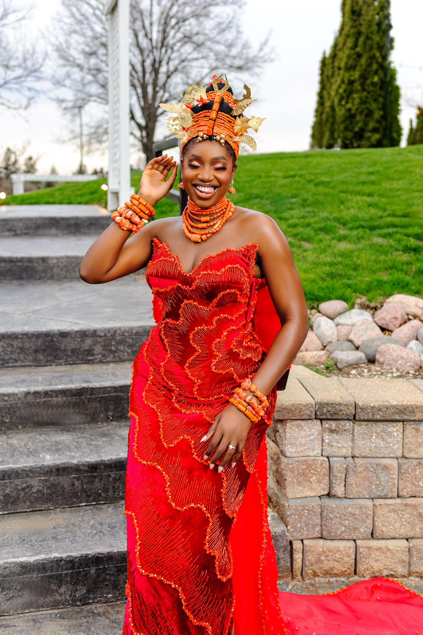Woman in vibrant red Nigerian traditional attire with coral beads and ornate headdress smiling on outdoor steps.