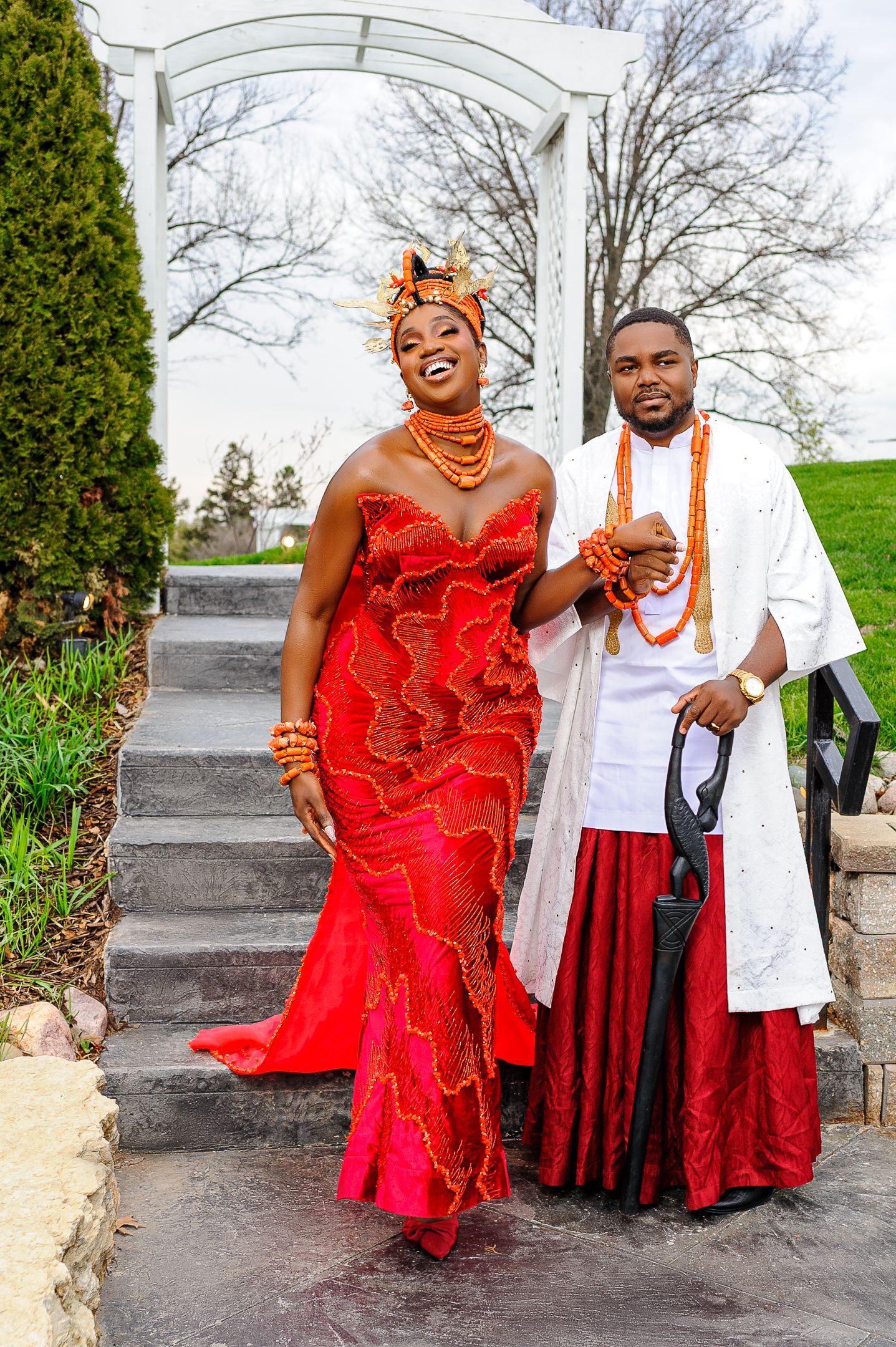Couple in vibrant Nigerian traditional wedding attire, woman in red gown, man in white agbada with coral beads.