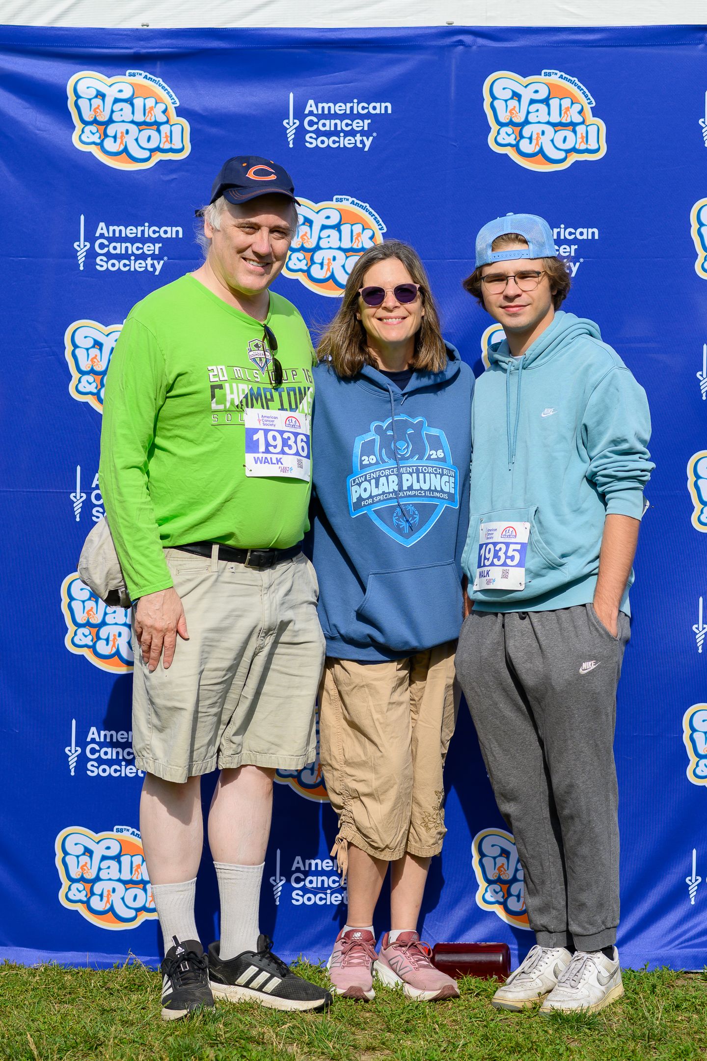 Three participants pose at American Cancer Society Walk & Roll event, wearing race bibs and casual athletic clothing outdoors.