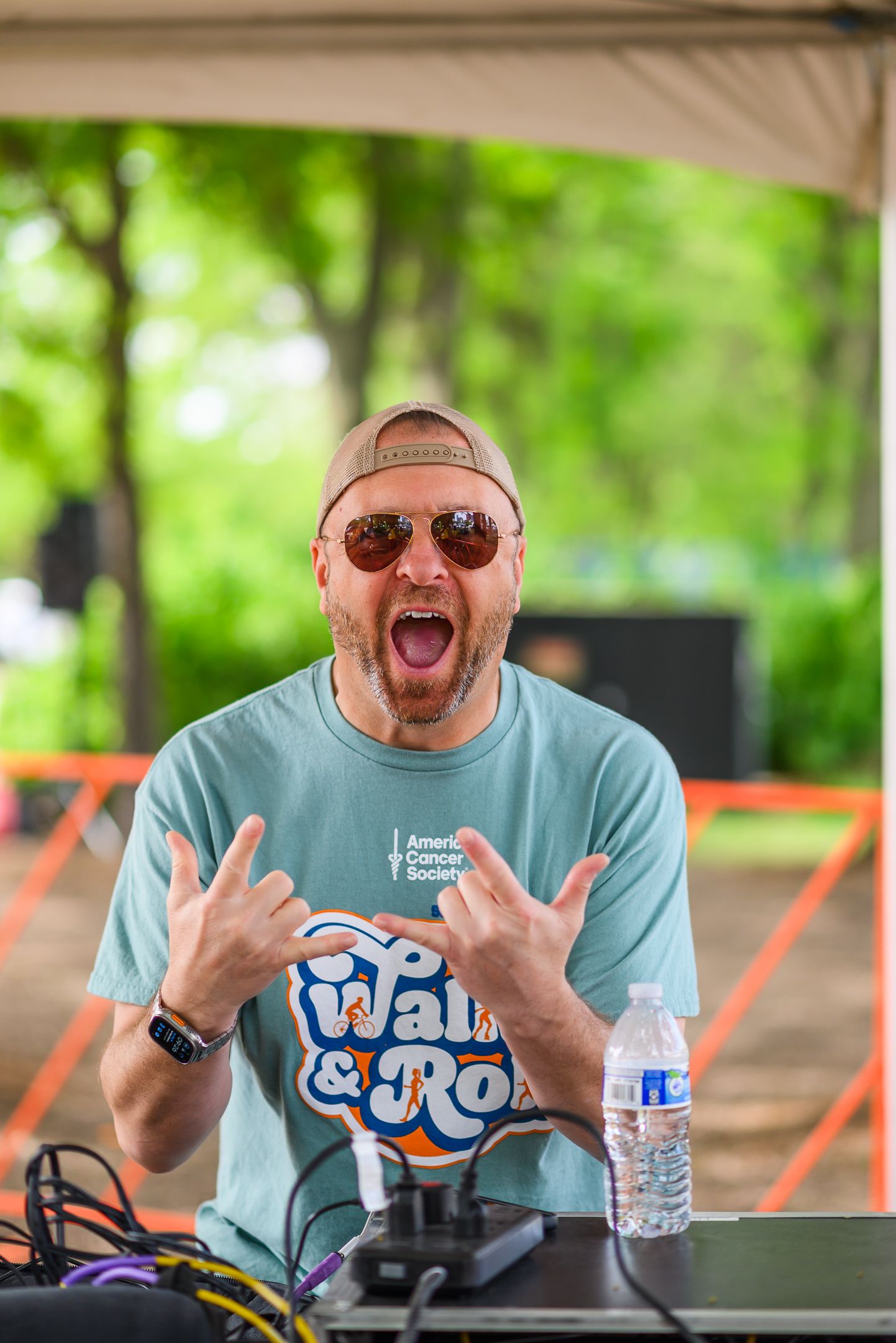 Energetic DJ in Walk & Roll American Cancer Society shirt makes rock hands gesture at outdoor event.