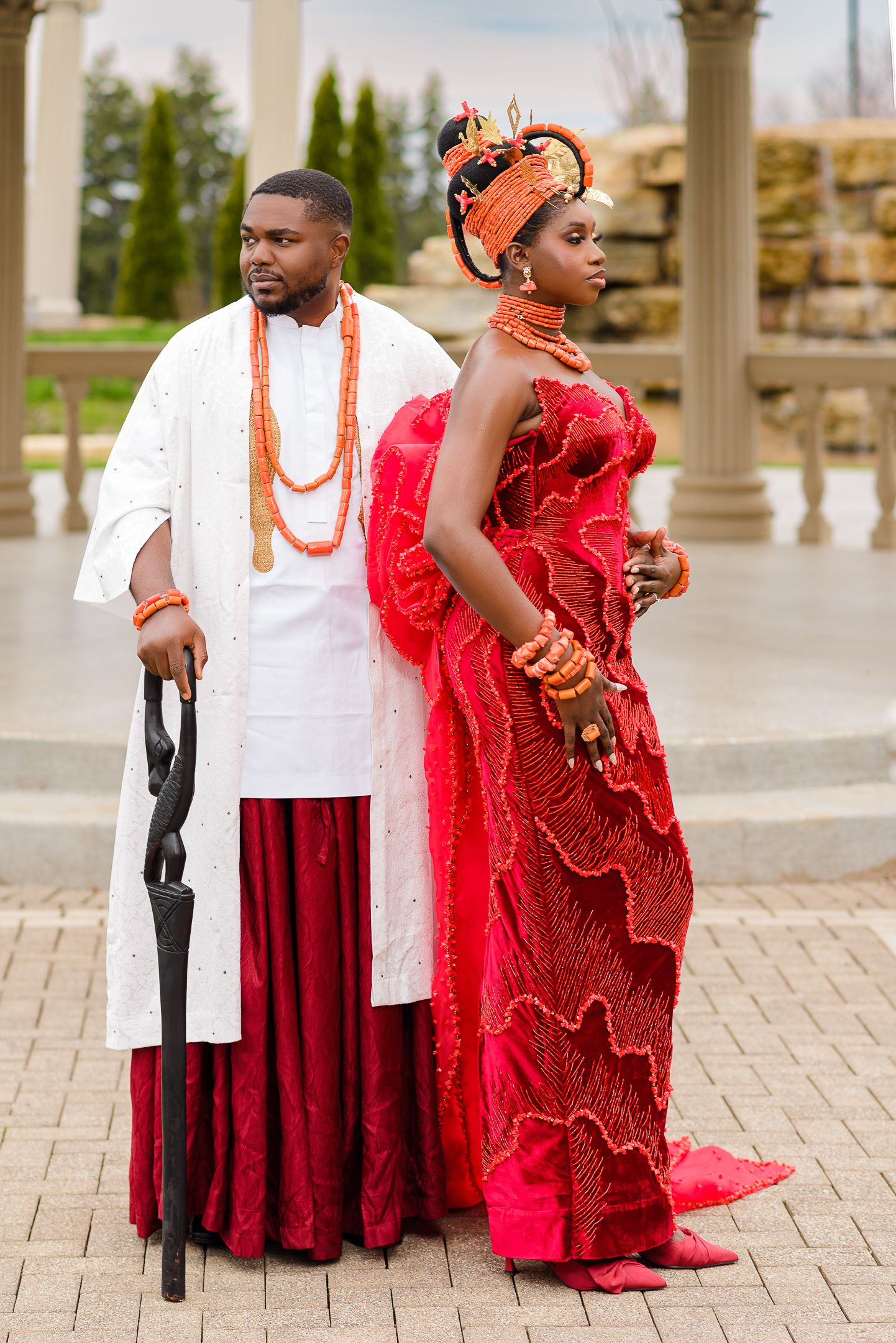 Couple in traditional Nigerian wedding attire, man in white agbada, woman in red lace gown with coral beads and headdress.