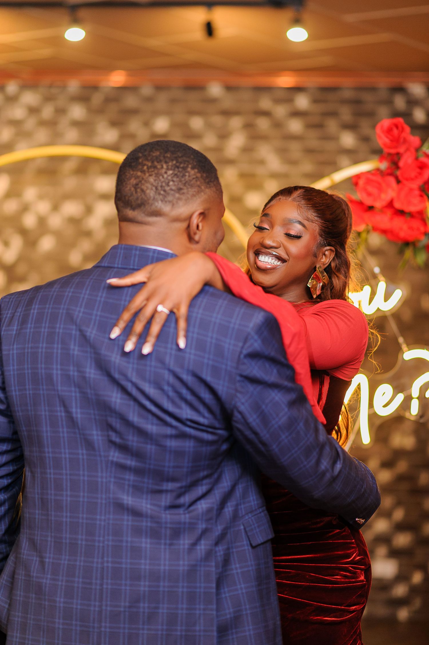 A joyful couple embraces during a romantic proposal, woman in red dress smiling broadly with roses and neon sign backdrop.