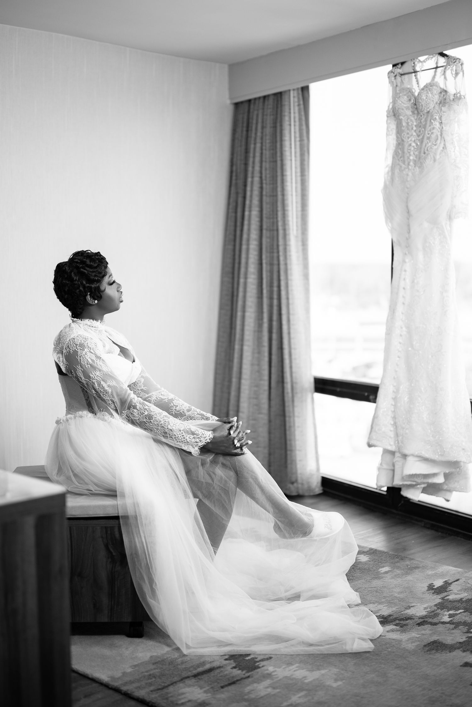 Bride in white lace gown sits contemplatively beside hanging wedding dress in black and white photo.