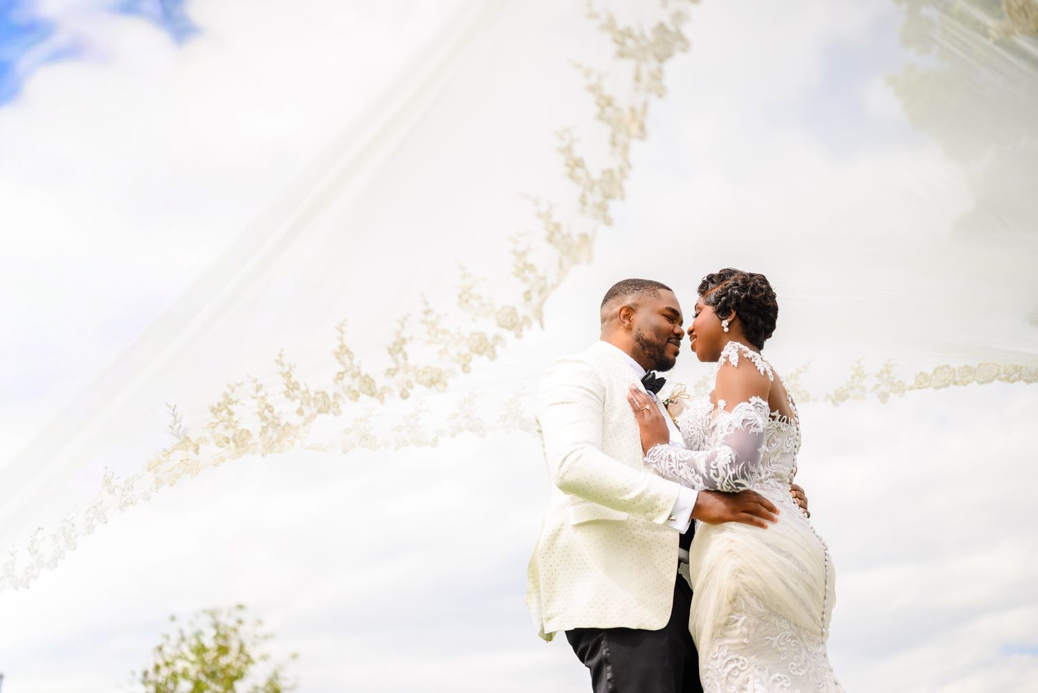 A bride and groom share a romantic embrace under a flowing white veil against a cloudy sky on their wedding day.