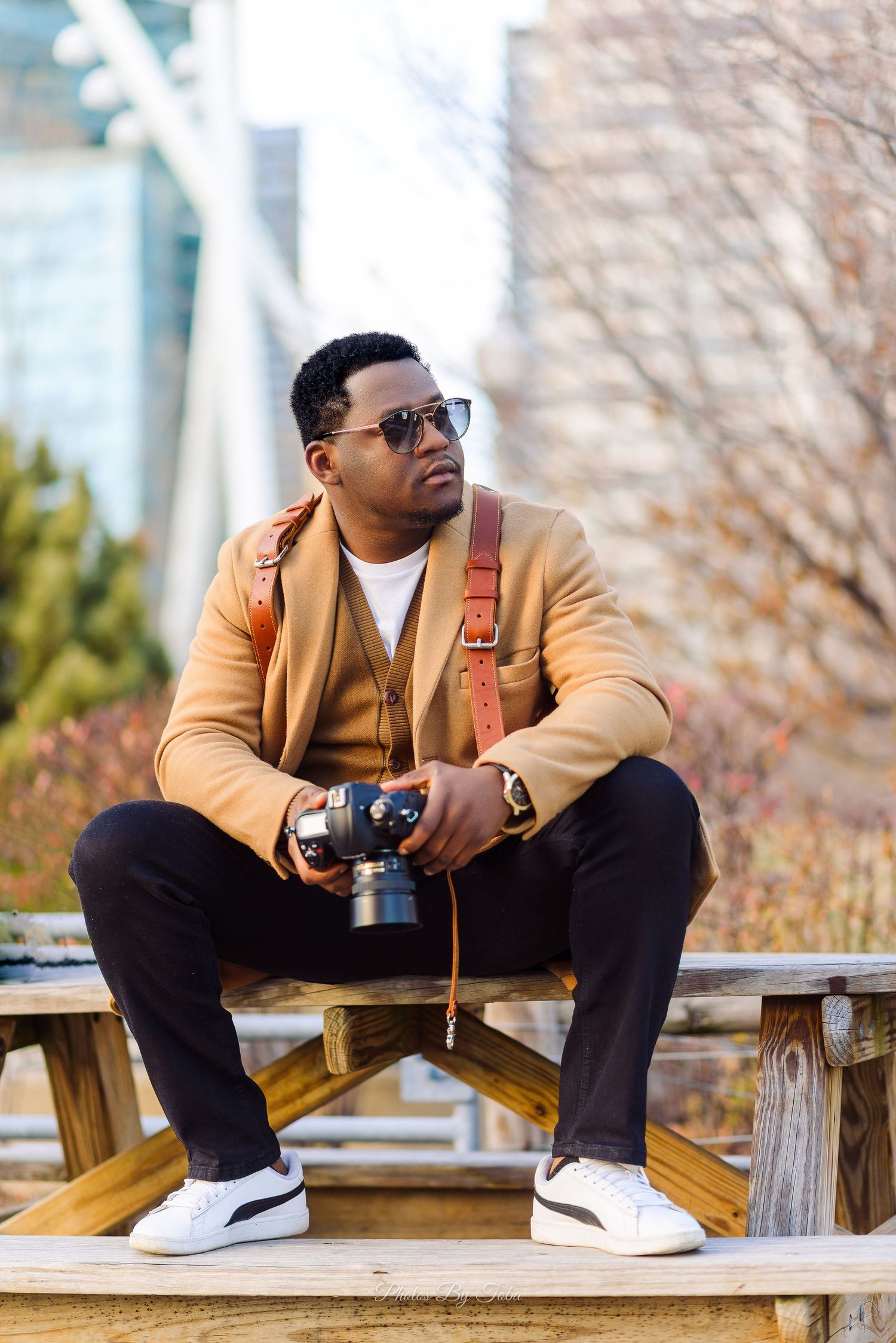 A stylish man in a tan blazer and sunglasses poses thoughtfully on a wooden bench in an outdoor setting.